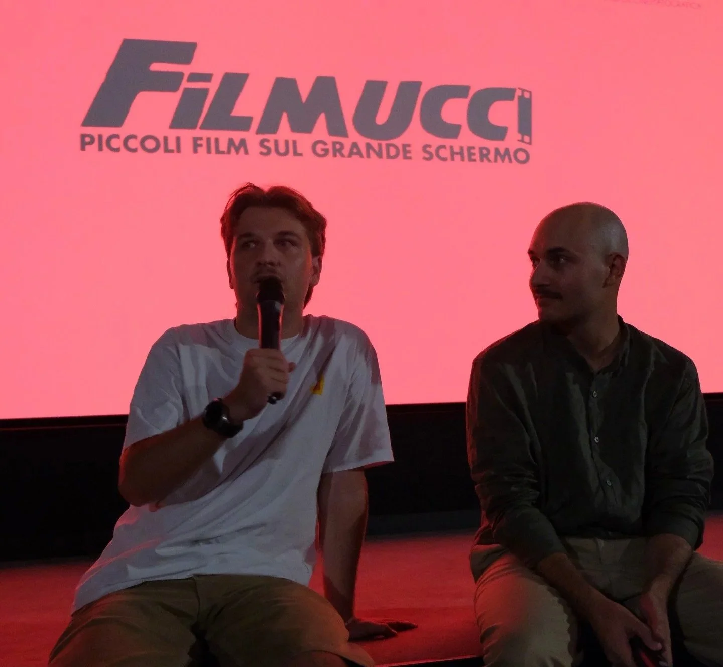 Two men sitting on stage in front of a red screen with the FilmMucc logo and Italian text, one speaking into a microphone while the other listens.