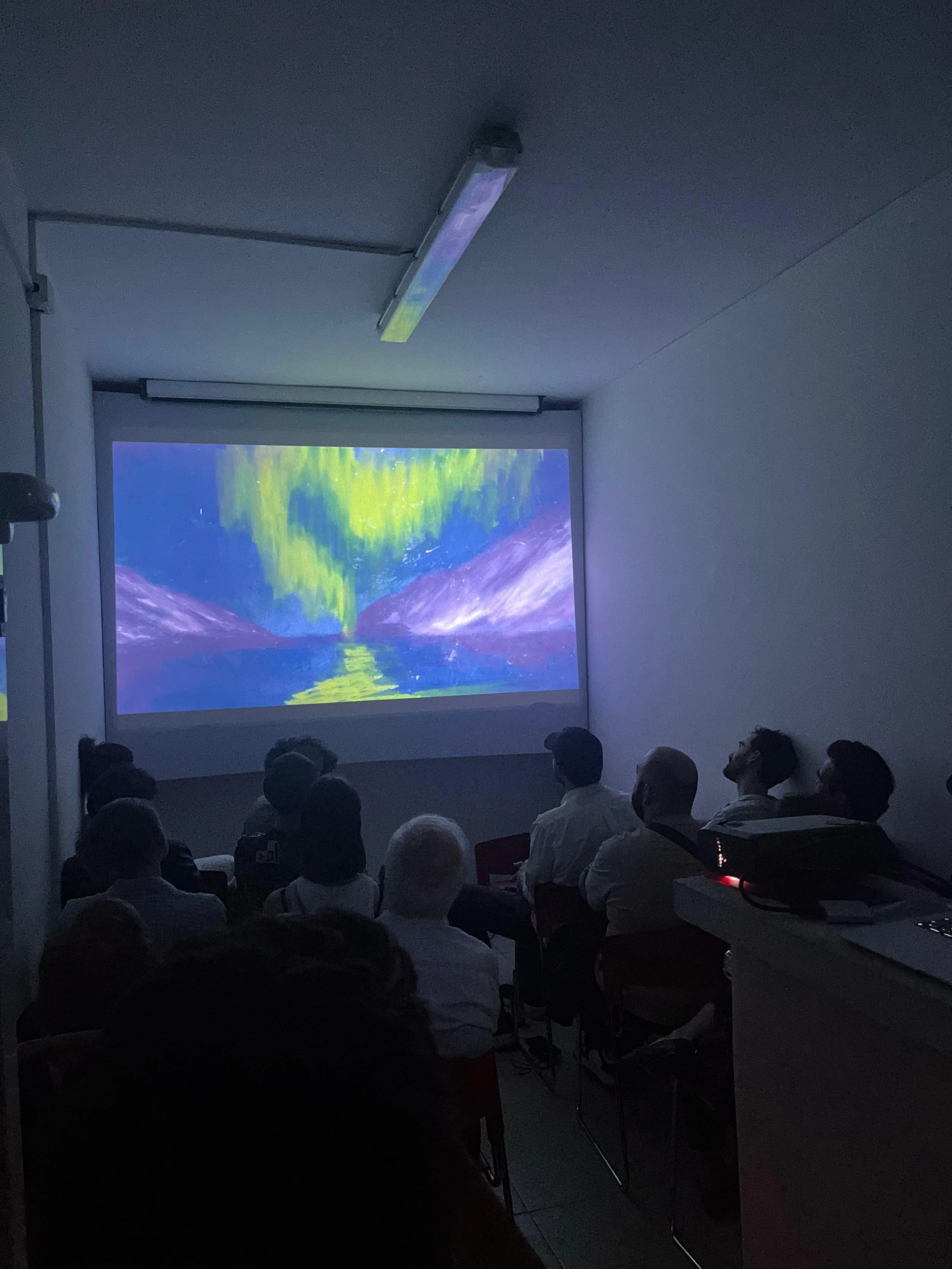 People sitting in a dark room watching a projection of the Northern Lights over mountains reflected in a lake.
