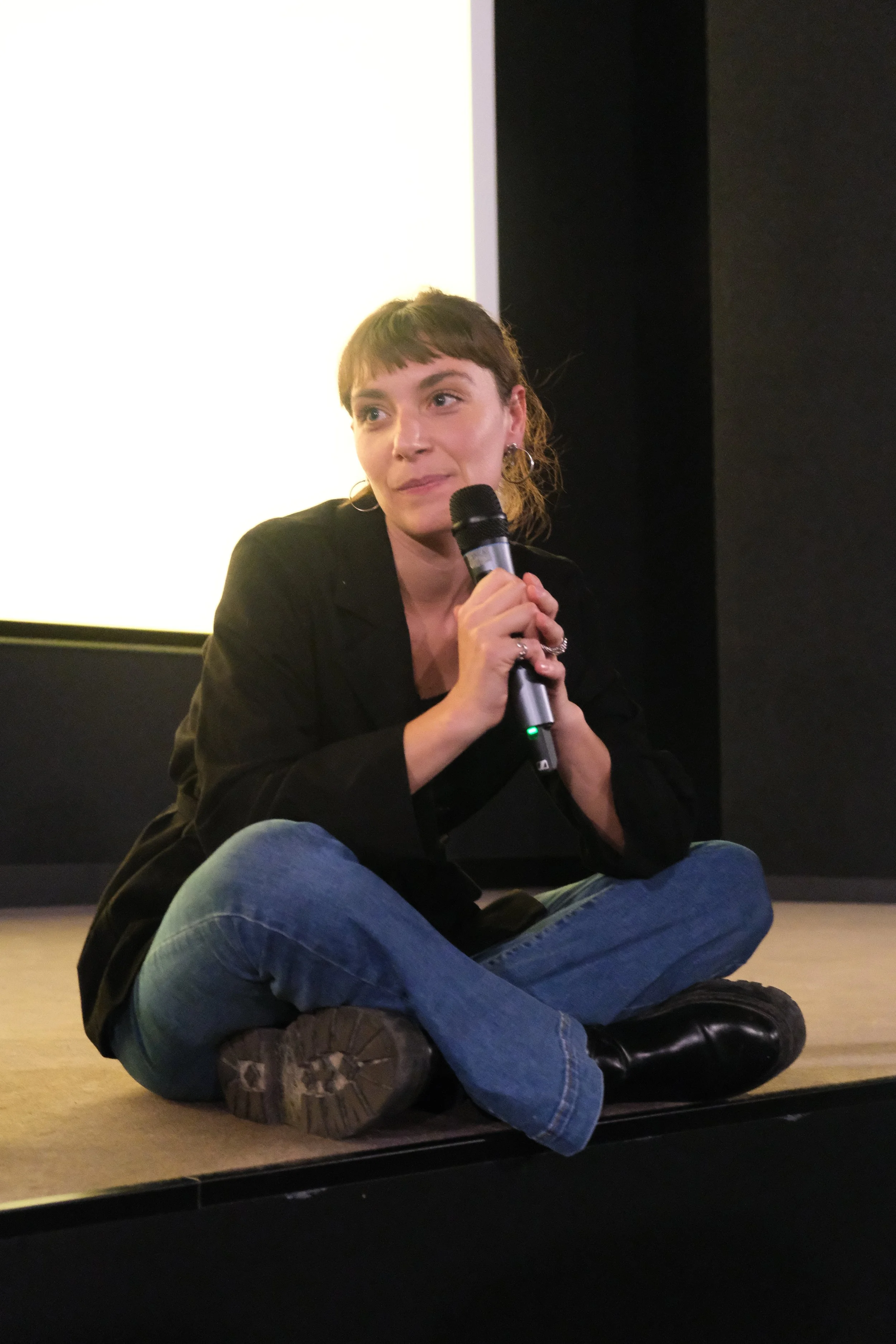 A woman sitting cross-legged on the floor, holding a microphone, speaking during a presentation or event.