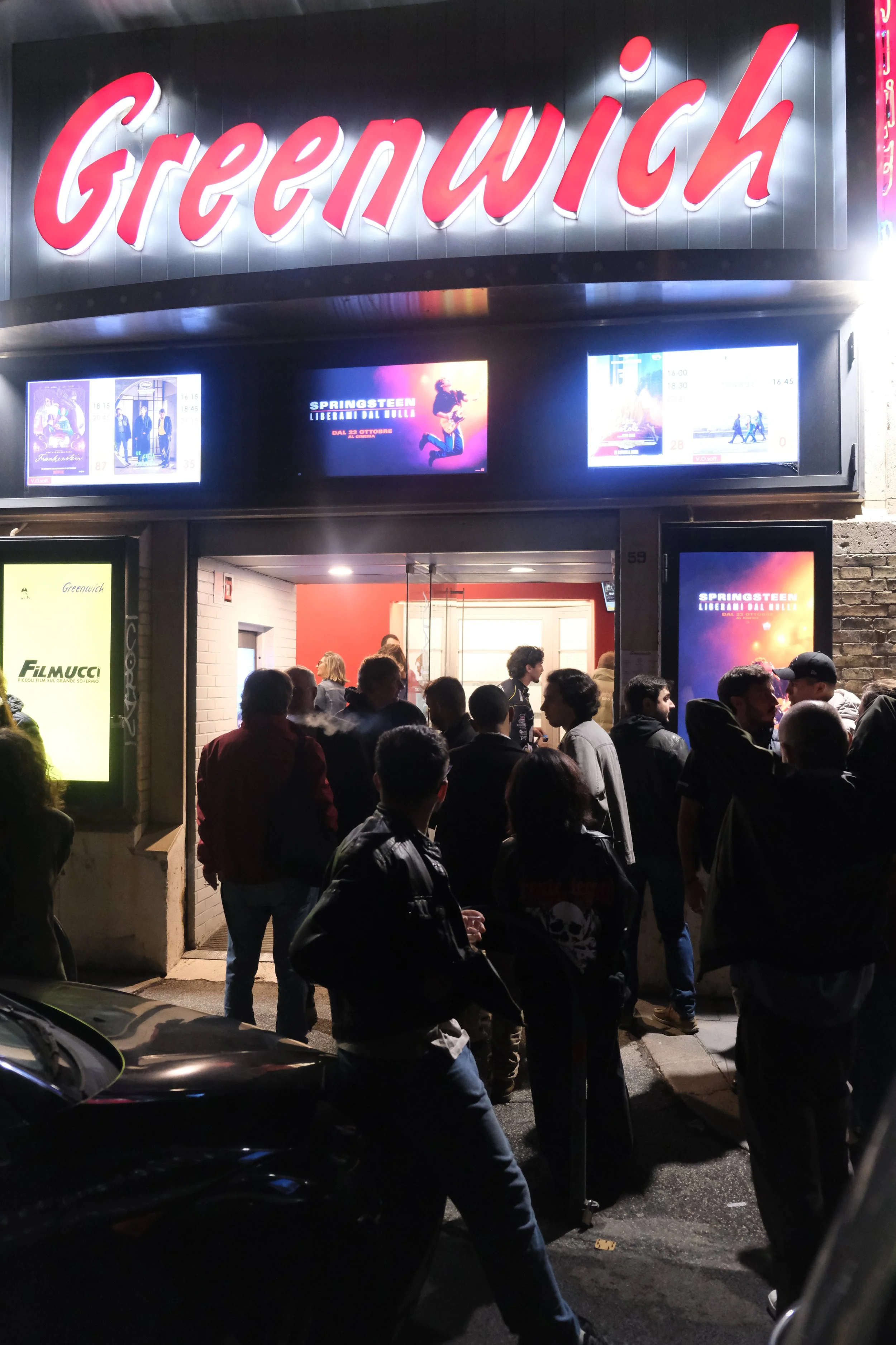People gathered outside the Greenwich cinema at night, with illuminated digital screens displaying movie advertisements and a neon sign reading 'Greenwich' above the entrance.