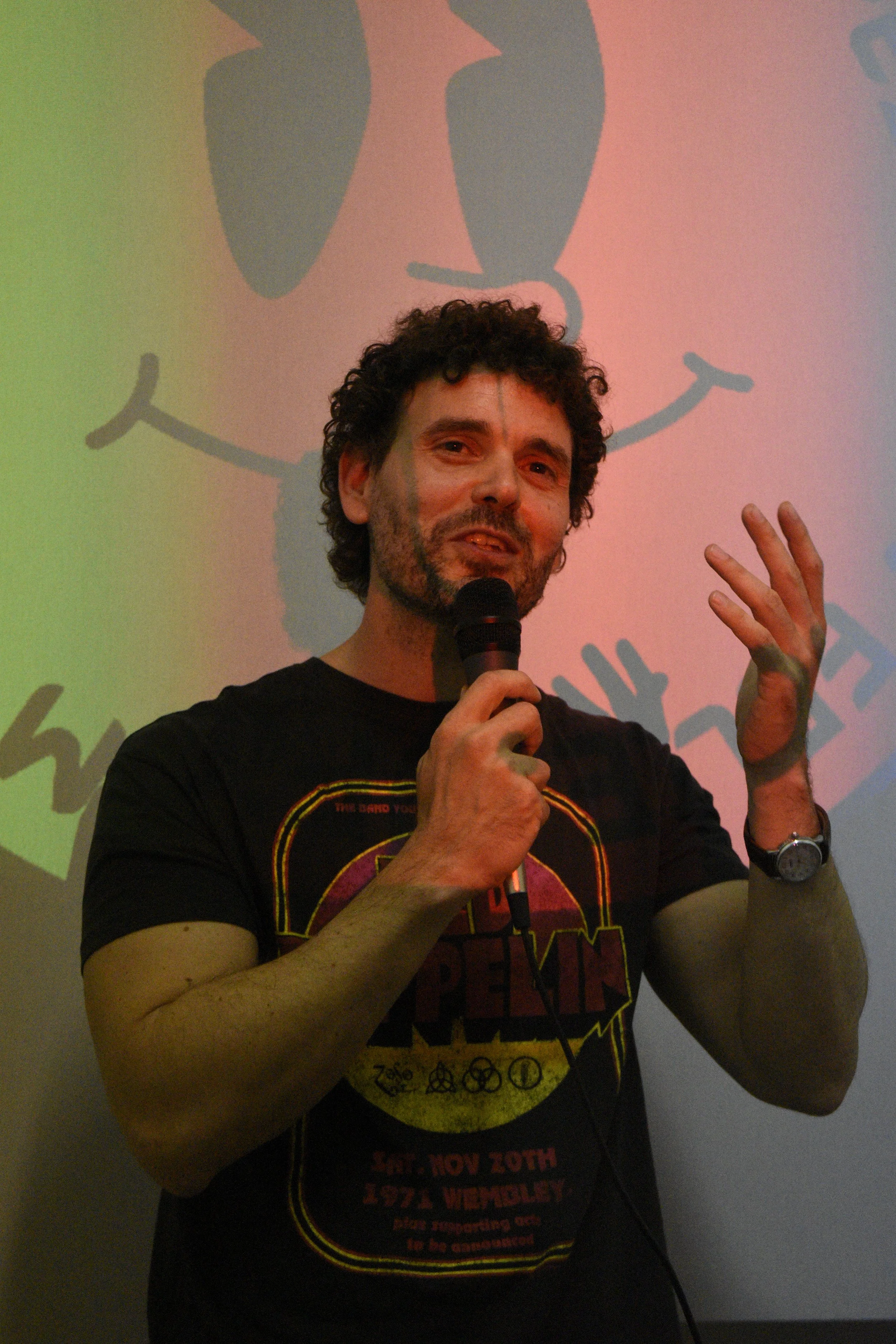 A man with curly hair holding a microphone and gesturing with his right hand, standing in front of a colorful background with a drawing of a dog.