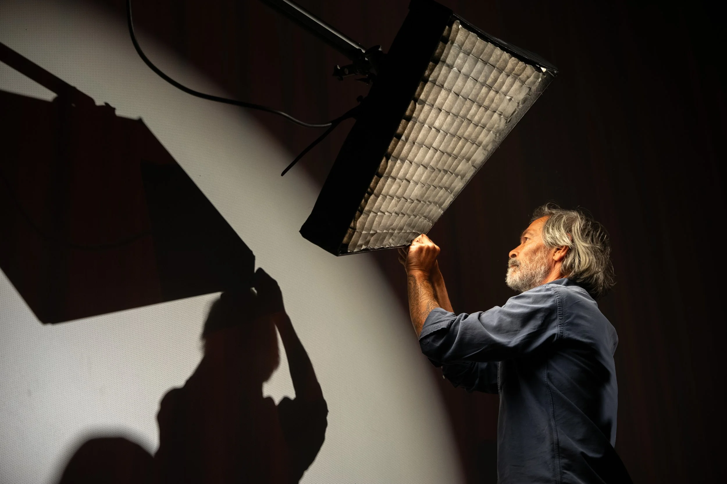 An older man with gray hair and a beard adjusts a lighting fixture against a wall, creating a shadowed silhouette.