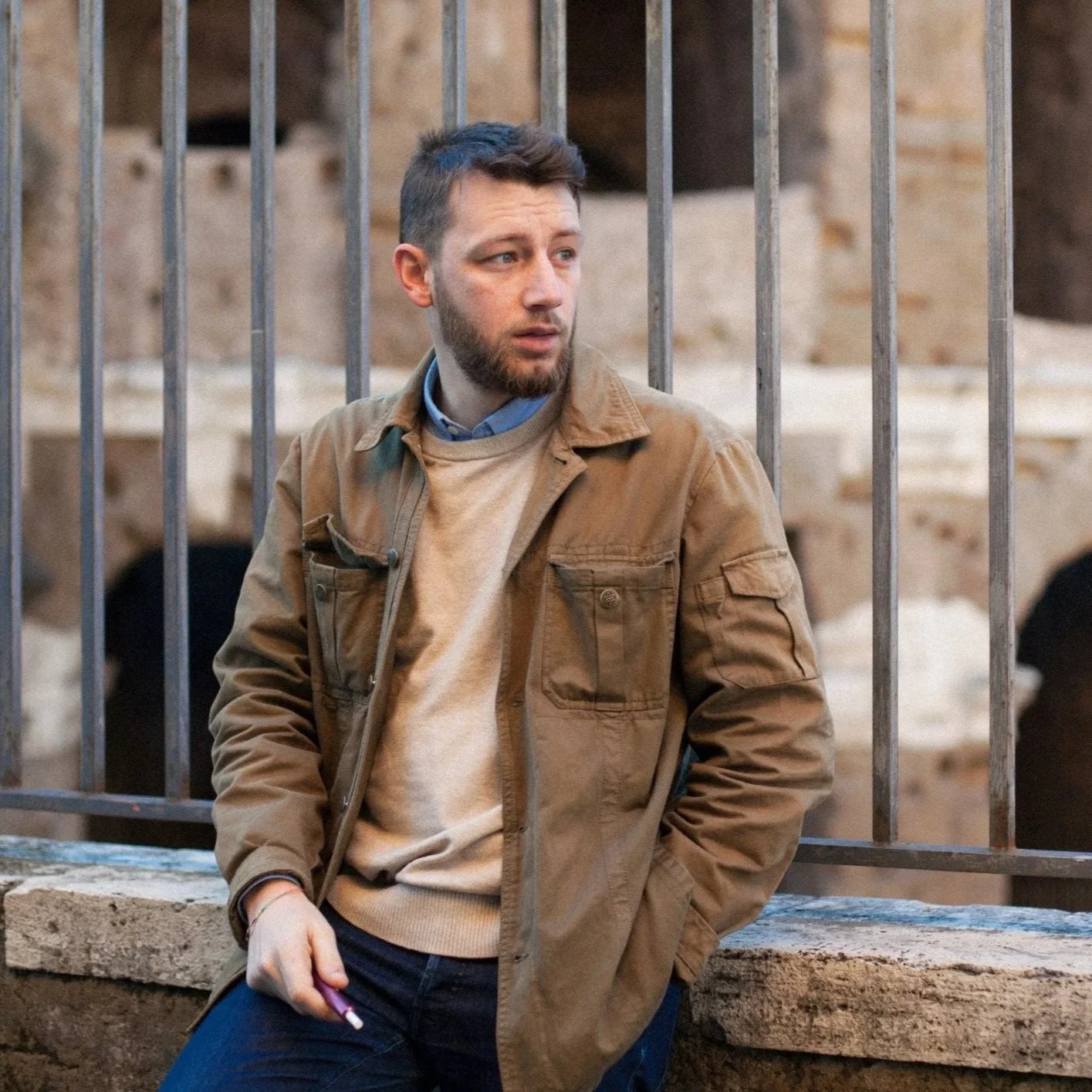 A young man with short brown hair and a beard standing outdoors near a metal fence, holding a cigarette in his right hand, wearing a tan jacket over a beige sweater and a blue collared shirt.