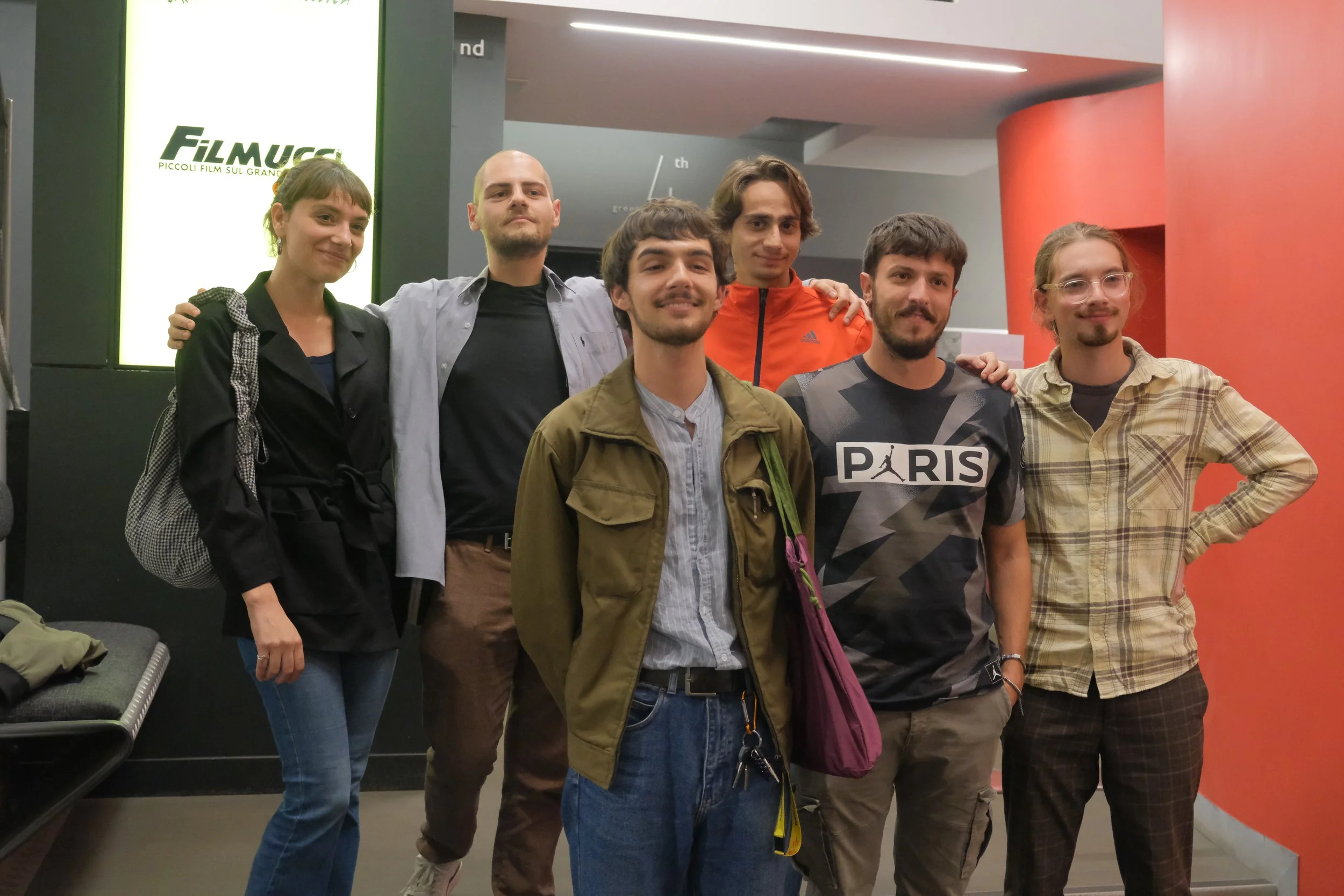 Group of seven young adults posing together at a cinema or theater lobby, standing in front of a bright sign that reads 'FilmUcca'.
