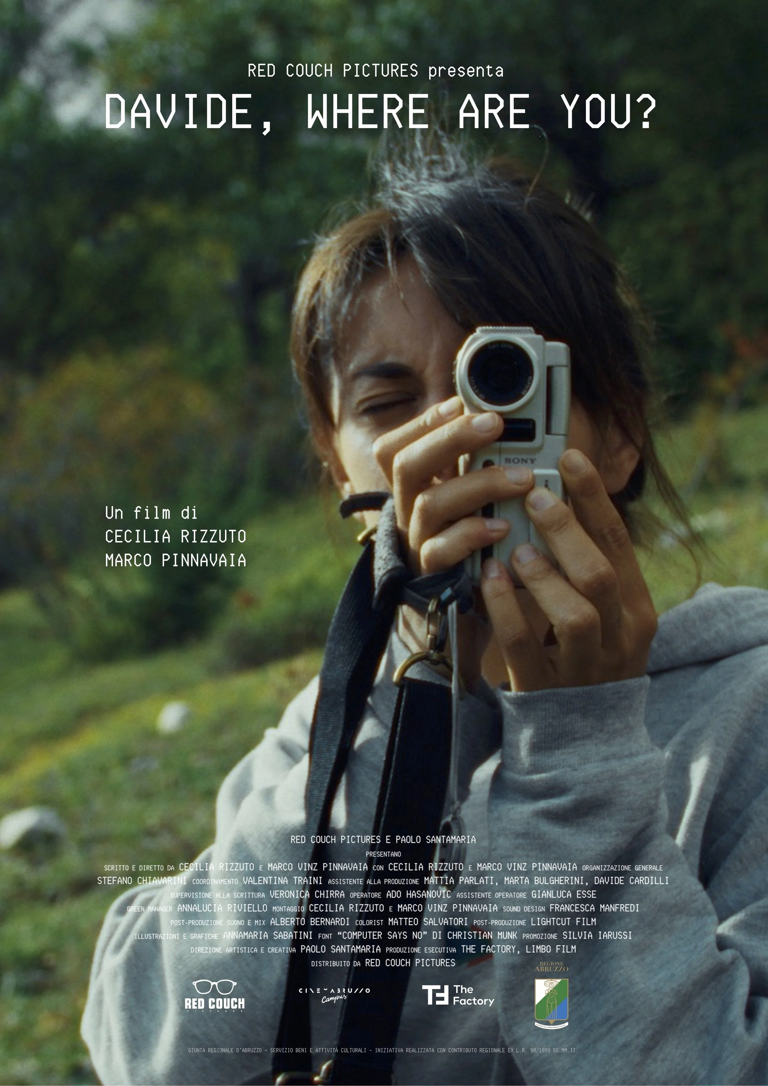 A person with dark hair is holding a vintage camera up to their face, taking a photo outdoors with greenery in the background. The person is wearing a gray hoodie.