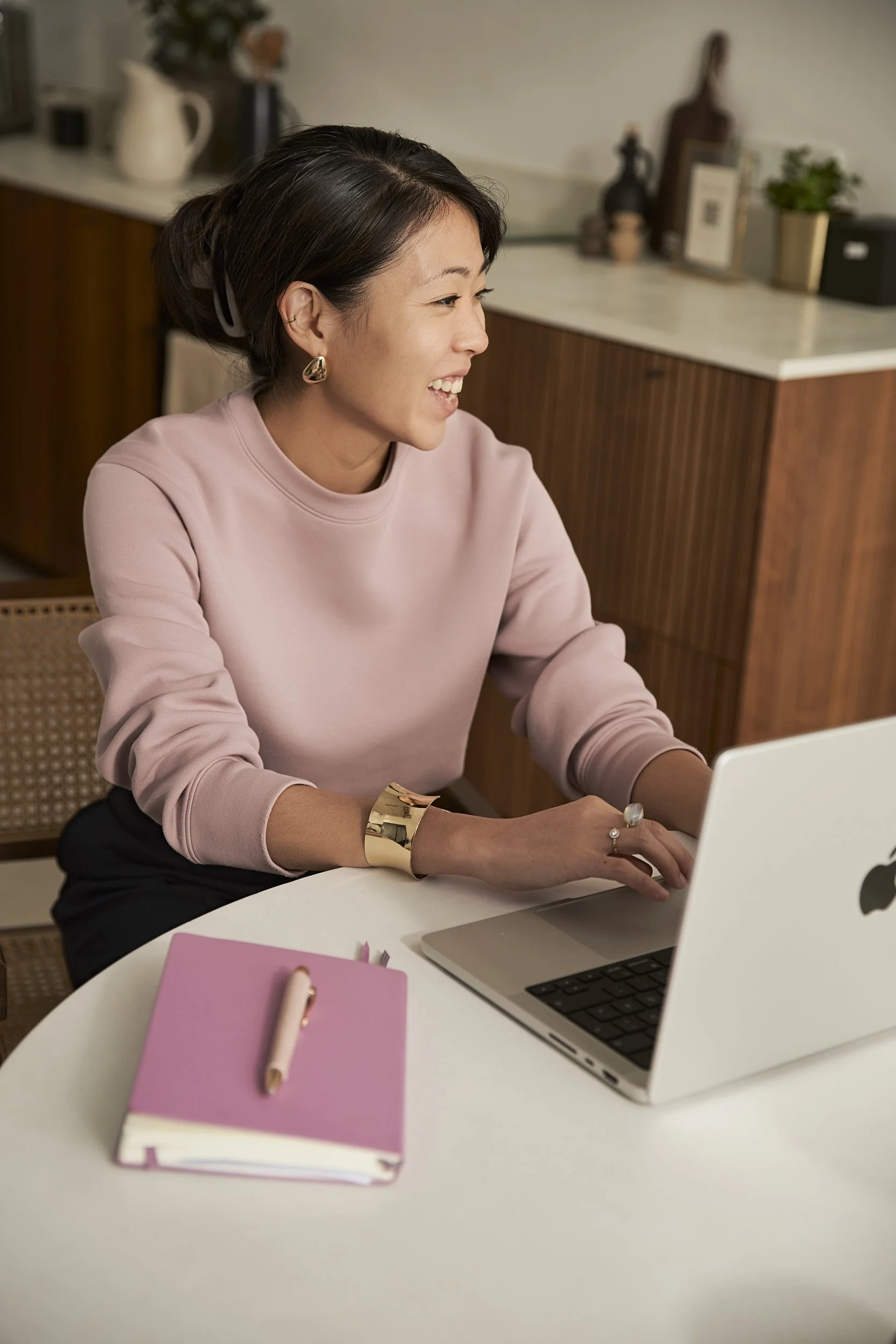 Eine lachende Frau arbeitet an einem Laptop in einer modernen Wohnung, neben einem pinken Notizbuch mit einem Stift.