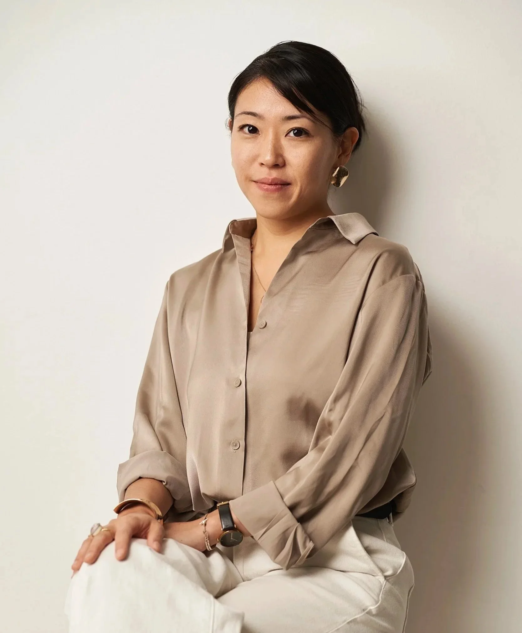 Ein Porträt einer Frau in einem beige Satin-Bluse, sitzend gegen eine helle Wand, mit kurzen dunklen Haaren, goldenen Ohrringen, Armbändern und einer Uhr.