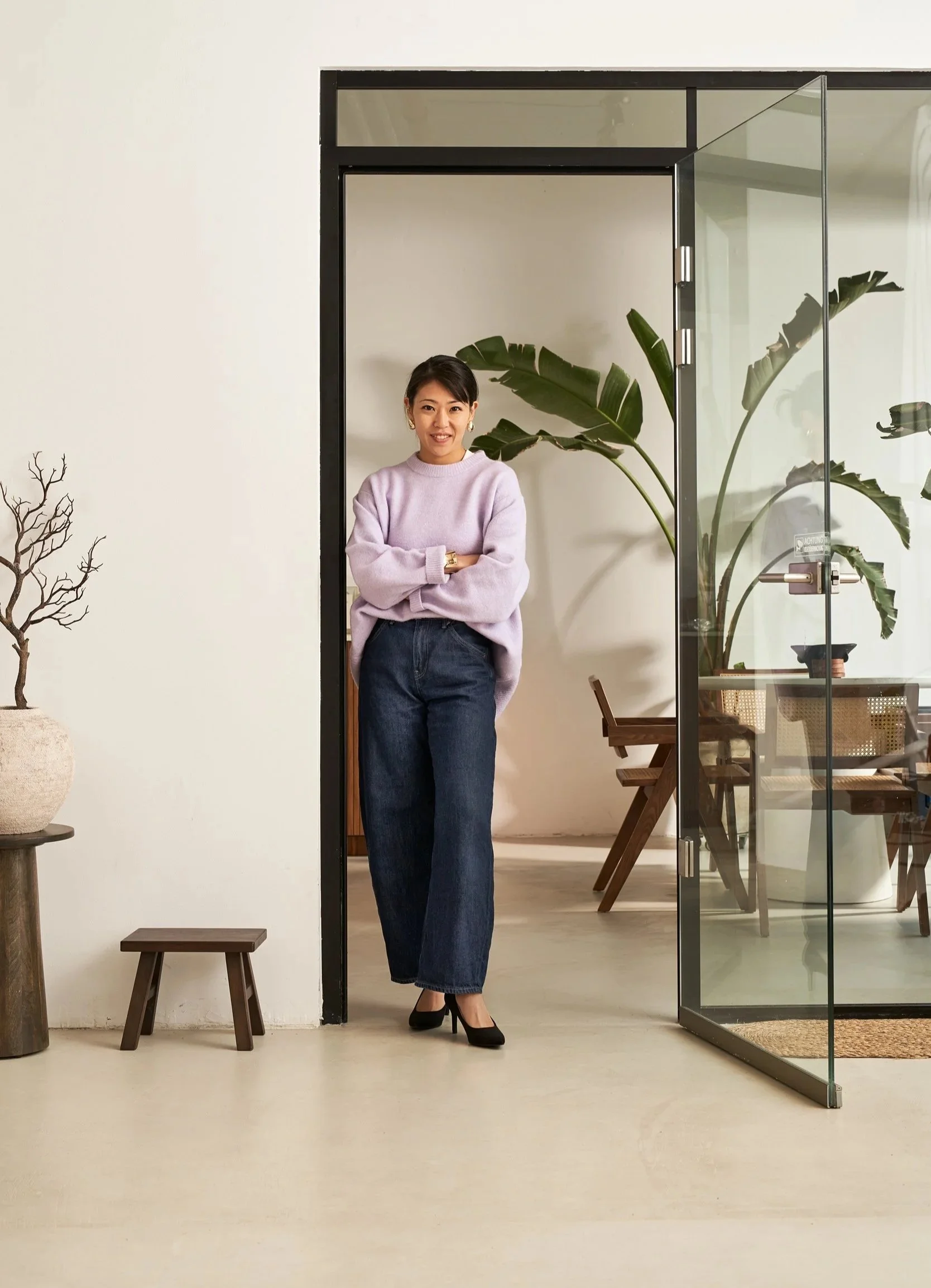 Eine junge Frau steht in einem modernen, hellen Raum, vor einem offenen Glas-Schiebetür mit großen Pflanzen im Hintergrund, trägt einen lilafarbenen Pullover, dunkle Jeans und schwarze High Heels.