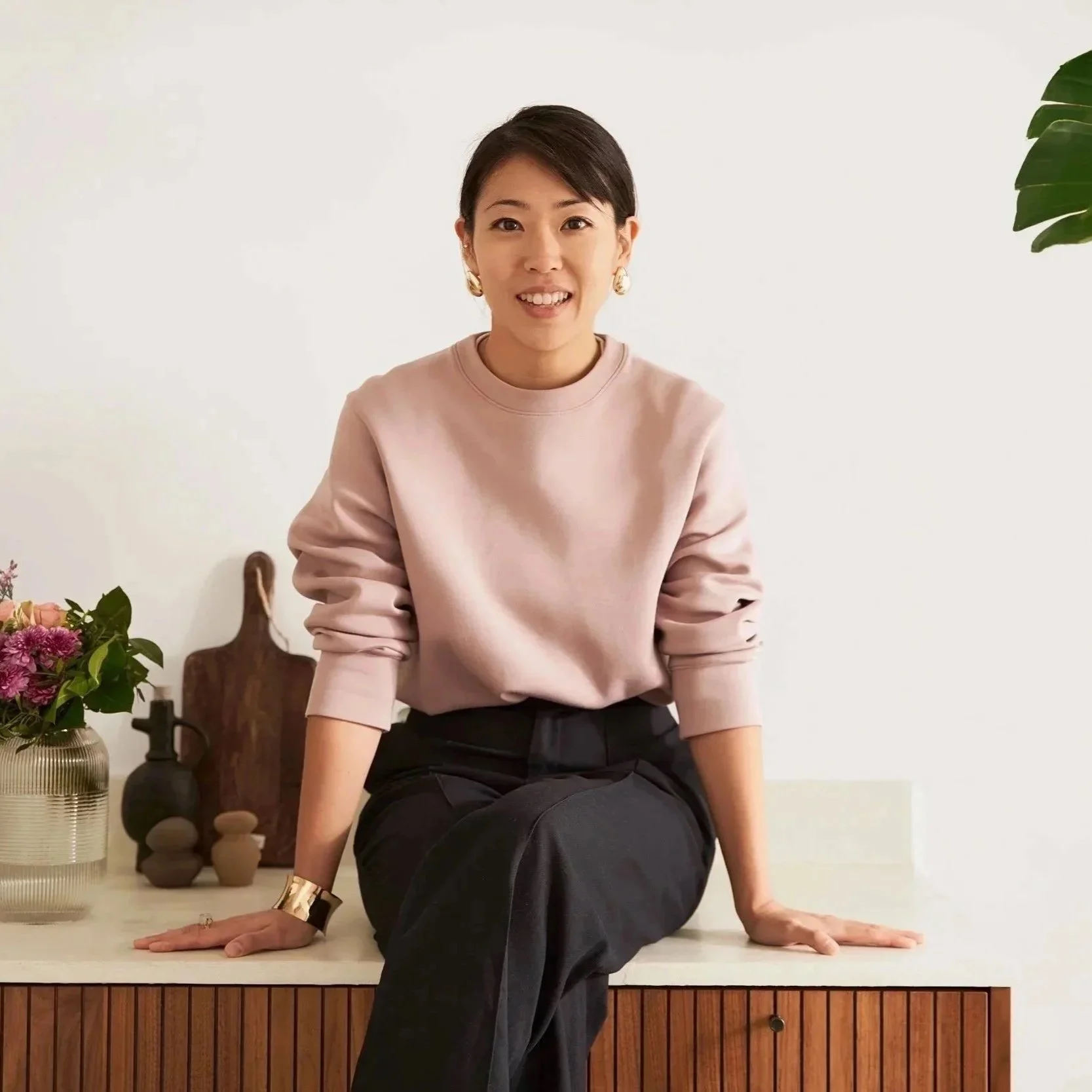 Eine junge Frau sitzt auf einer weißen Oberfläche vor einer weißen Wand, neben einer Vase mit Blumen und Dekorationsgegenständen, trägt ein rosa Sweatshirt und schwarze Hosen, lächelt in die Kamera.