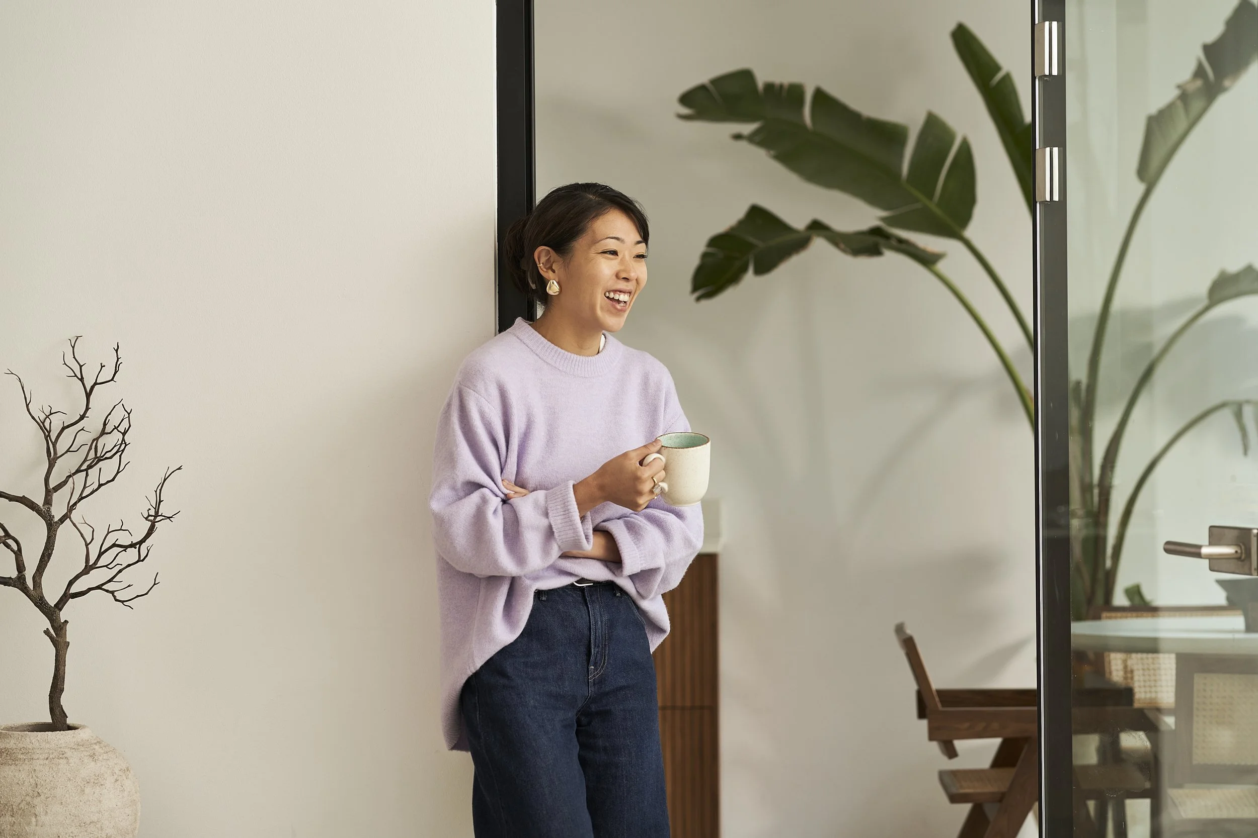 Eine lachende Frau im lila Pullover und dunklen Jeans hält eine Tasse und steht in einem modernen Raum mit weißen Wänden, großen Pflanzen und Glastüren.