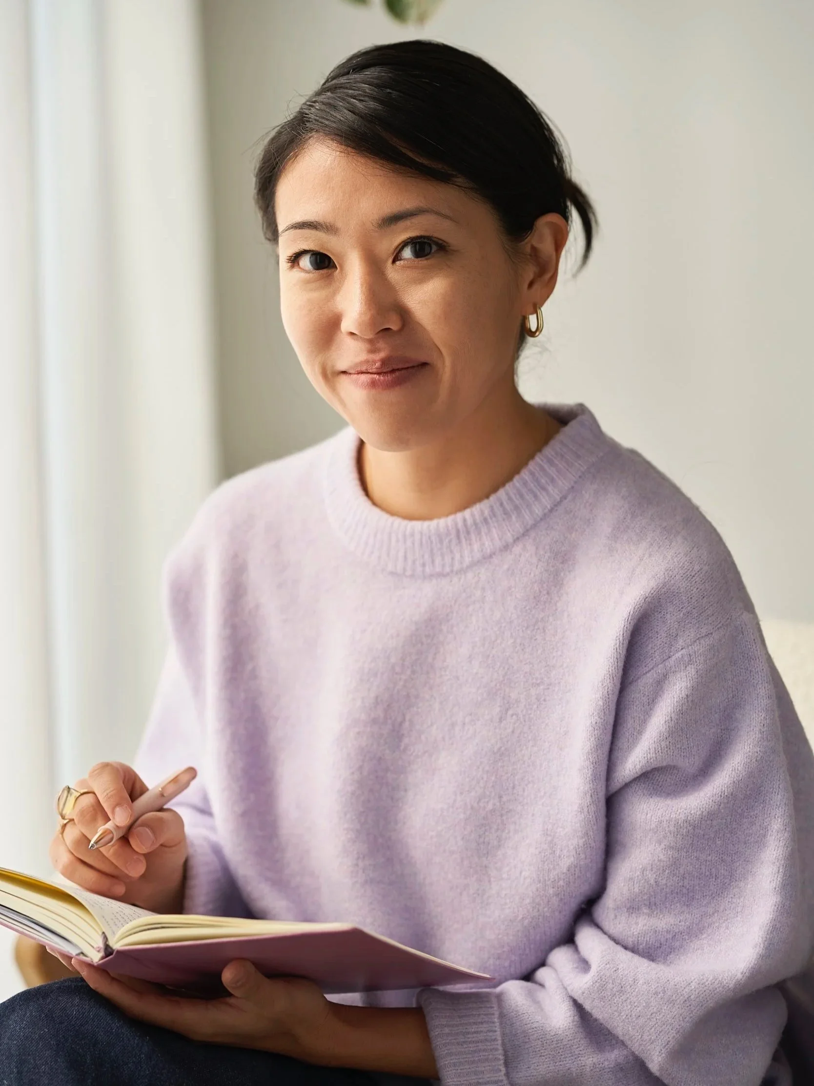 Eine Frau mit schwarzen Haaren, die einen lilafarbenen Pullover trägt, sitzt und schreibt in einem Notizbuch, vor einem hellen Hintergrund.