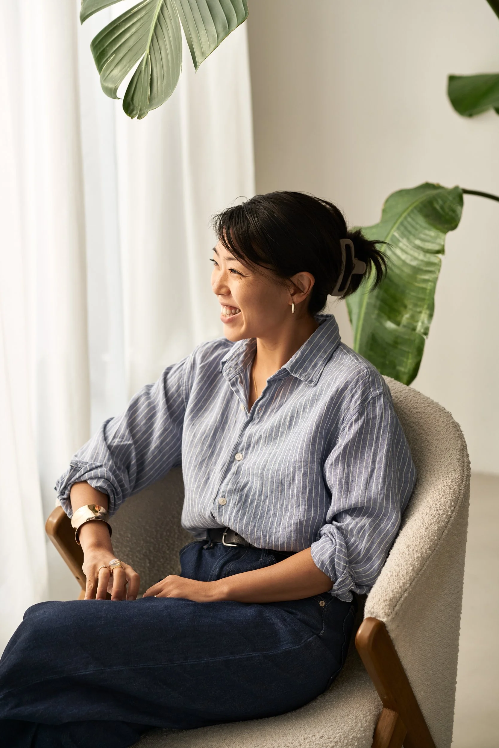 Frau sitzt nah am Fenster, lächelt, trägt einen blauen gestreiften Hemd, dunkle Hose und hat einen großen Haarclip, um die Haare zurückzuhalten. Es gibt große grüne Pflanzen im Hintergrund.