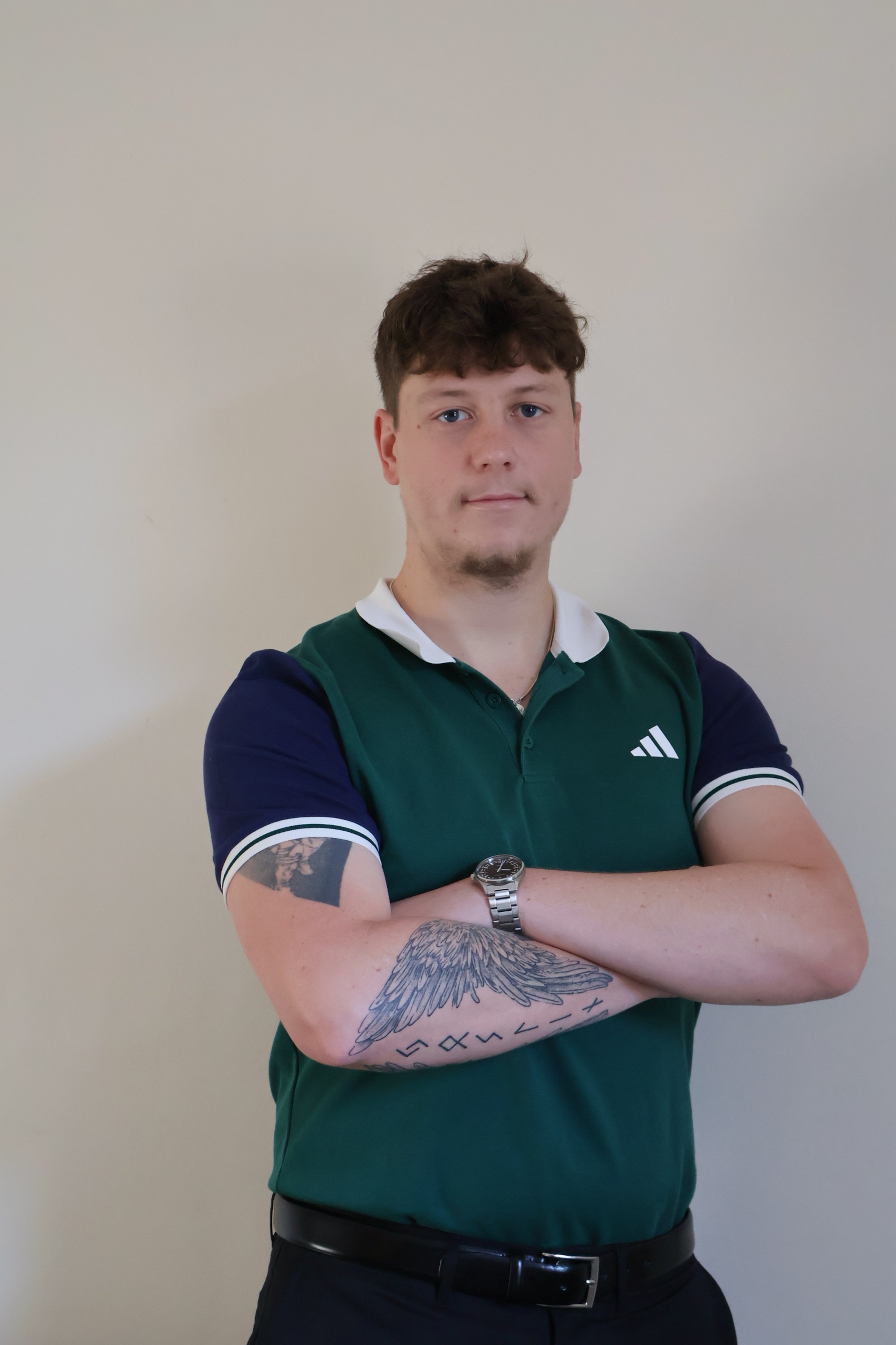 A physiotherapist with short brown hair, wearing a green and navy sports shirt with a white collar, crossing his arms, with tattoos of wings and symbols on his left arm, standing against a plain beige wall.