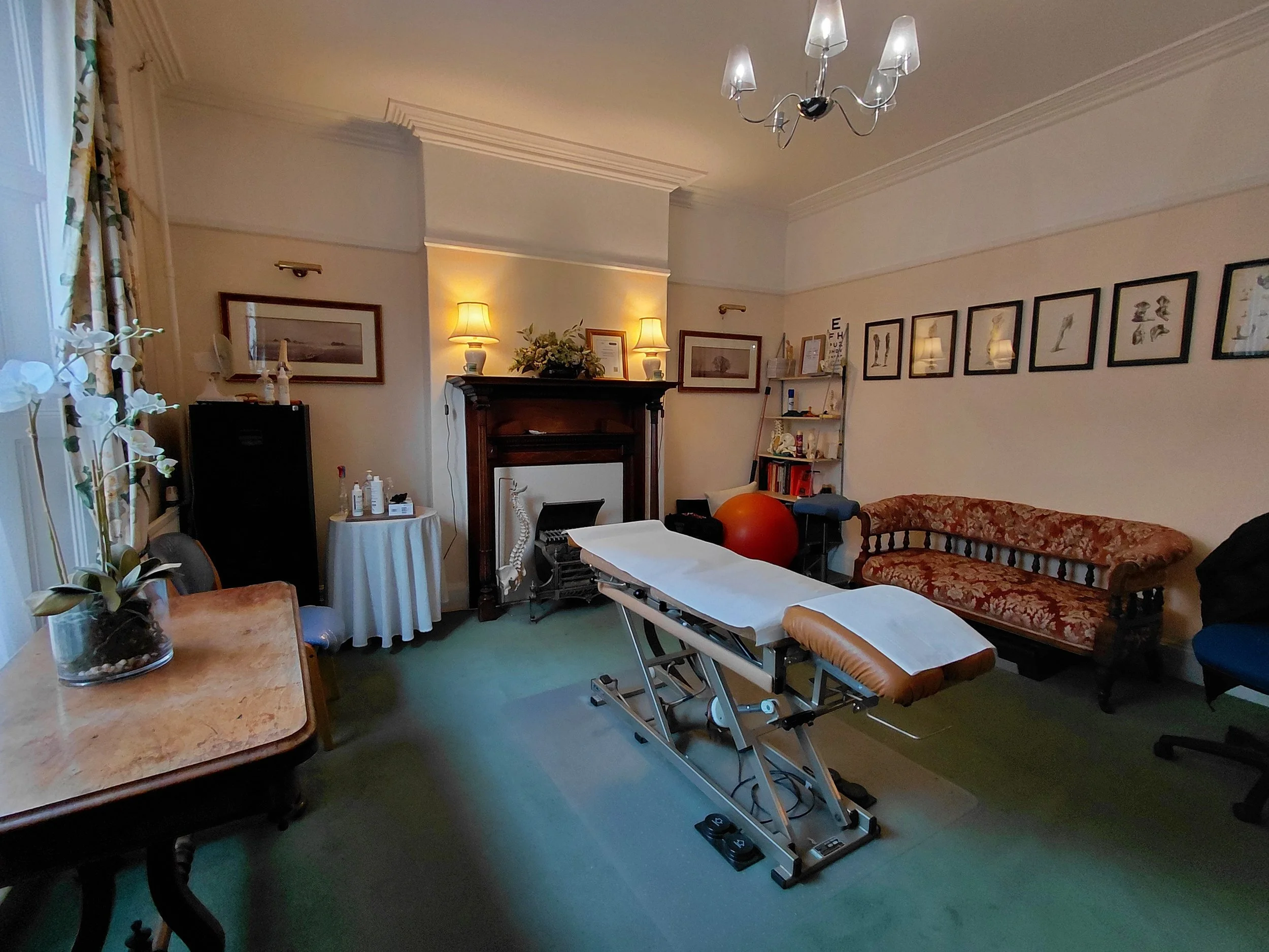 A room set up for a medical or therapy session, featuring a massage or examination table in the center, a vintage sofa against the wall, framed artwork and photographs on the walls, a small bookshelf, a large exercise ball, a desk with a potted orchid, and warm lighting from lamps and a chandelier.