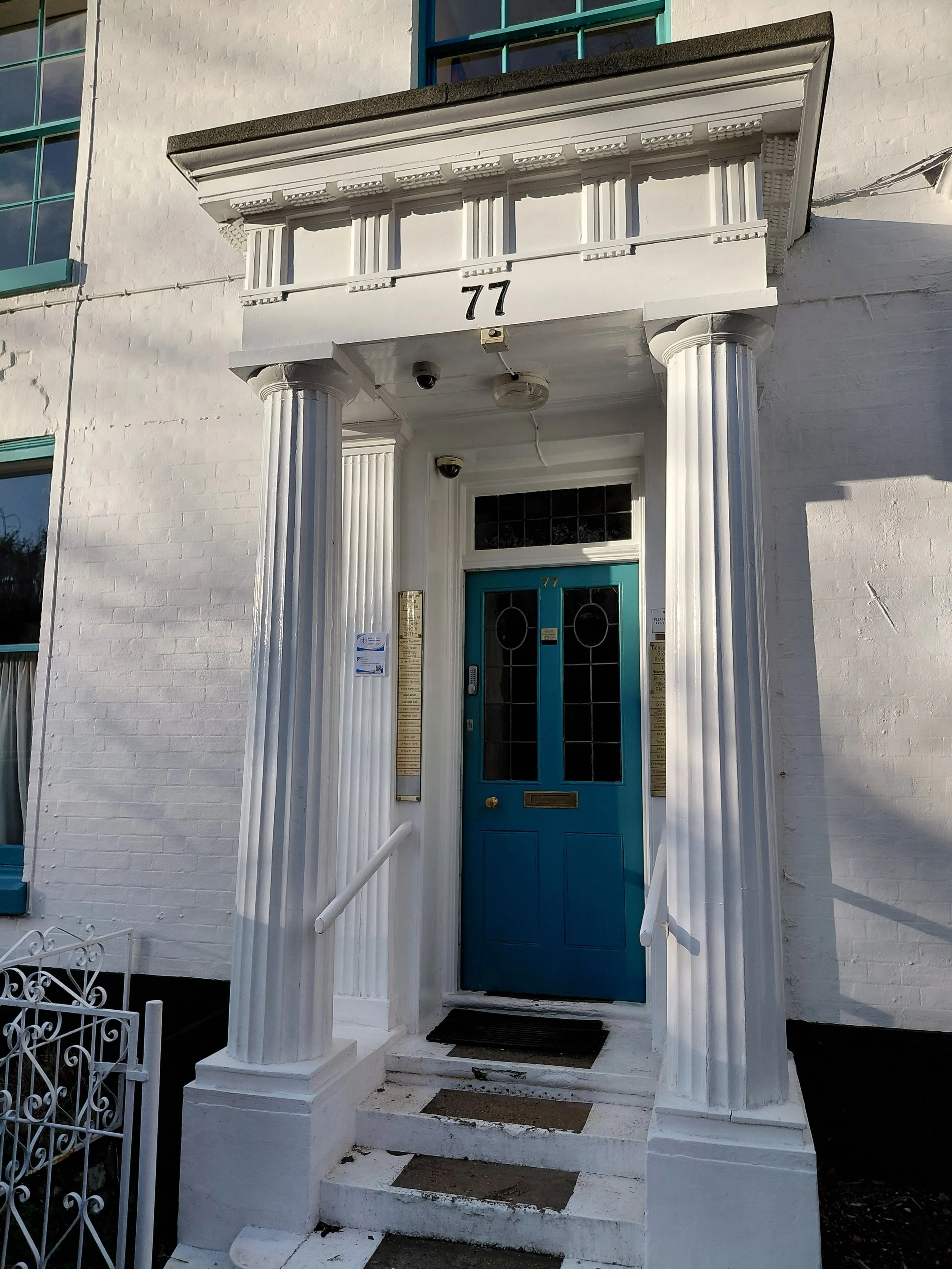 White brick building featuring a blue front door and large white columns at the entrance. The building number 77 is displayed above the door and on the balcony railing.