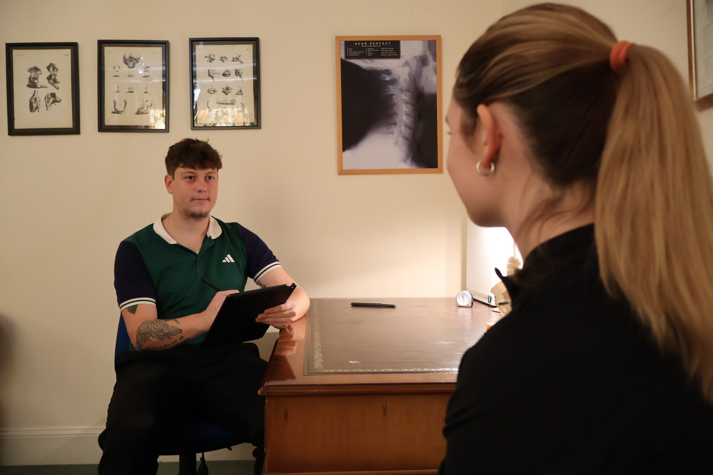A physiotherapist sitting at a desk during an interview or consultation with a patient. The physio is holding a tablet or clipboard and the patient is facing him, partially visible from the side. The room has framed diagrams and an X-ray on the wall.