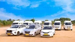 Six white vans and two white cars parked on a dirt lot under a blue sky with trees in the background.