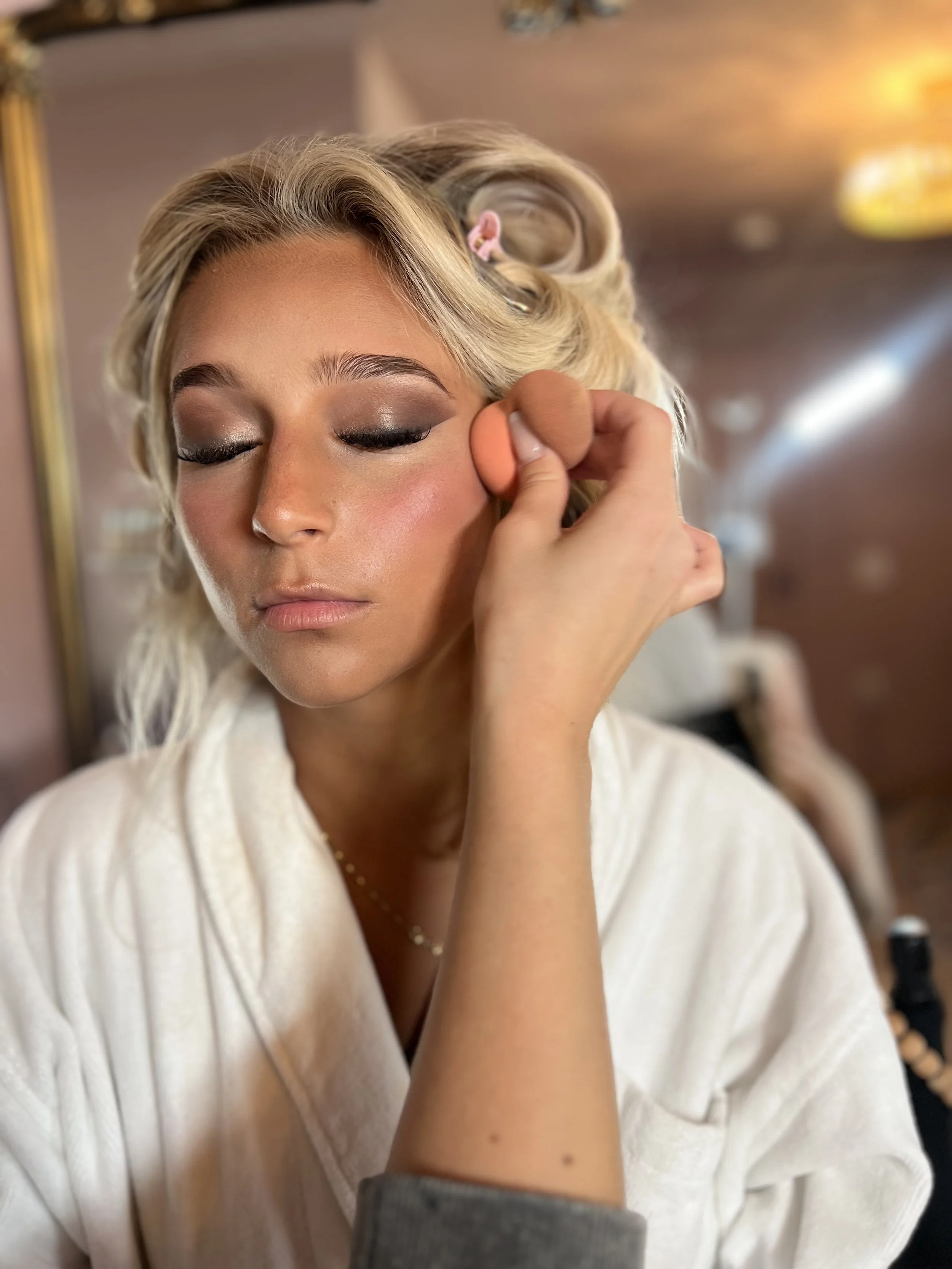 A woman with blonde hair and makeup having her face with a beauty sponge applied by a makeup artist.