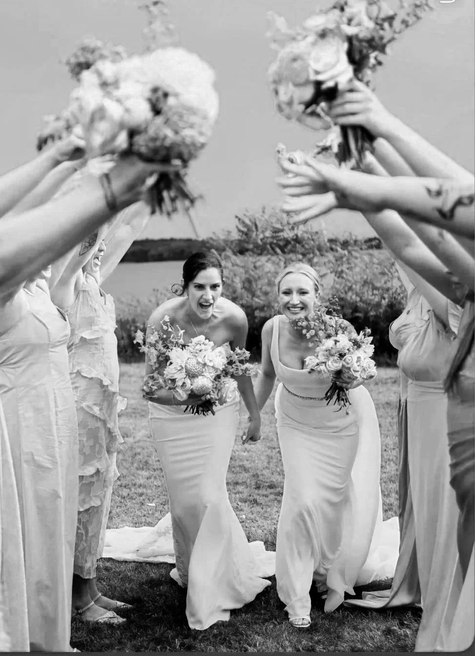 Two women in wedding dresses holding wedding bouquets walk through a tunnel formed by friends raising their bouquets in celebration, outdoors with water and trees in the background.