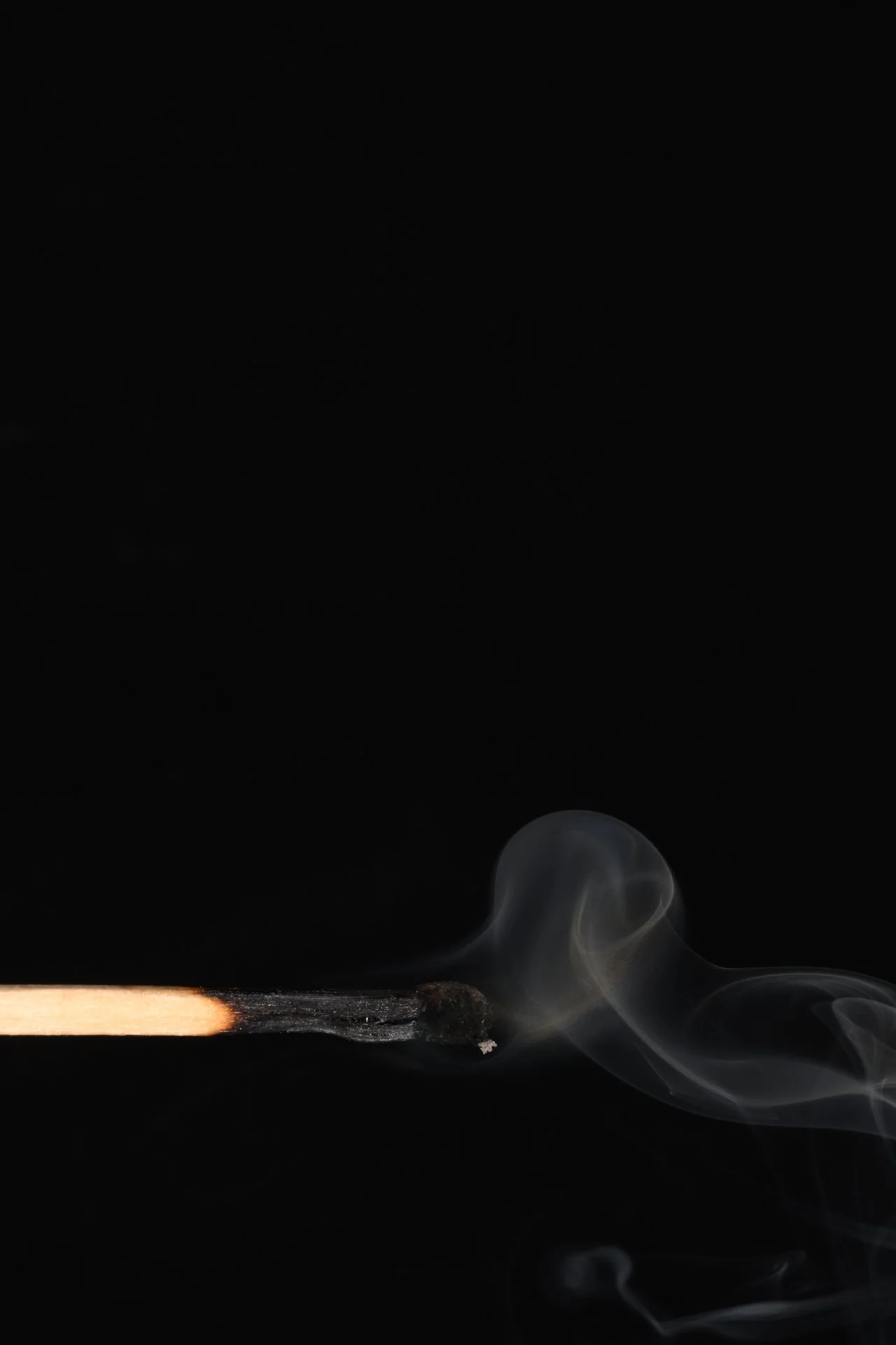 A lit matchstick with a glowing tip and wisps of smoke against a black background.