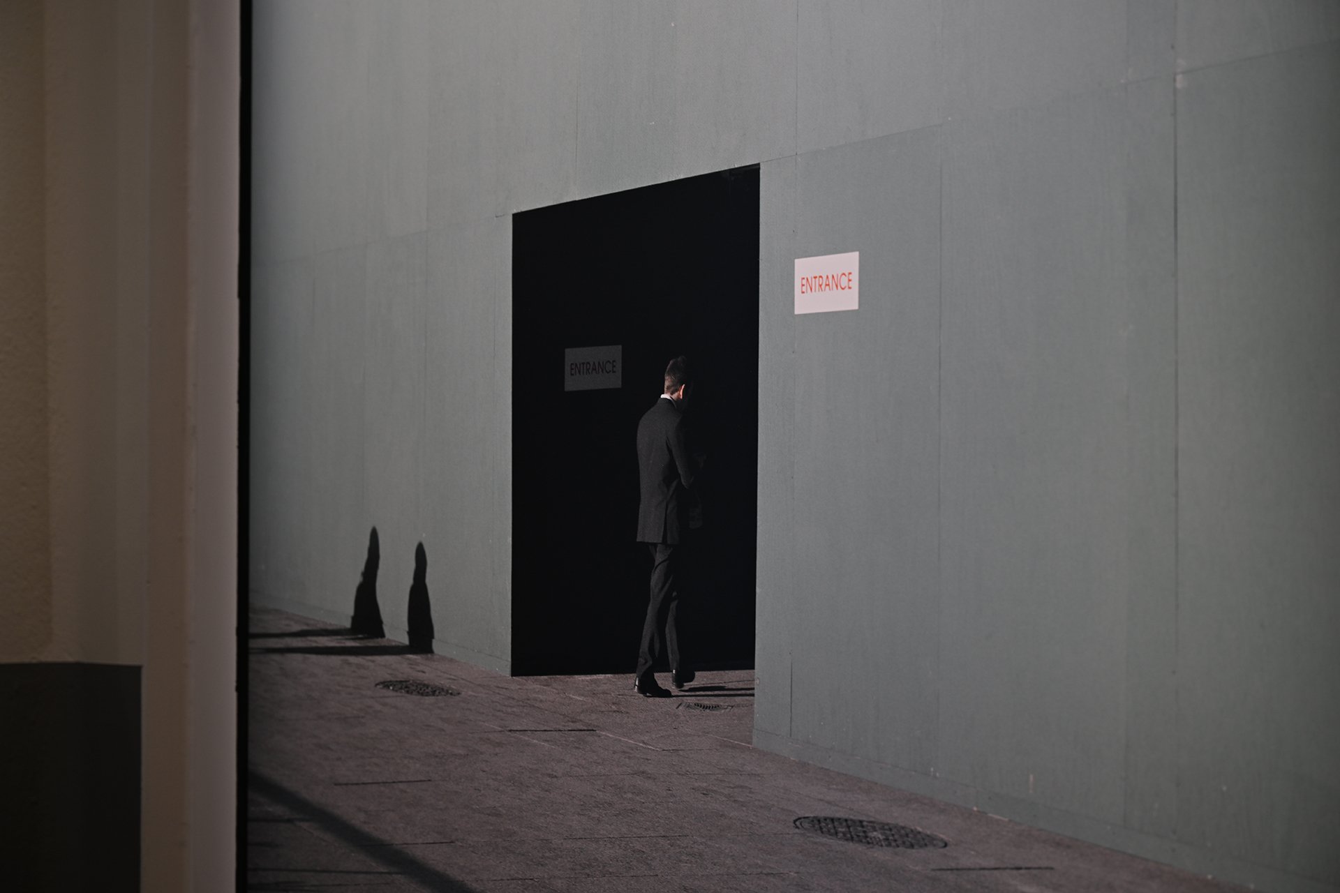 A man in a black suit stands outside an entrance to a building, with two signs that say 'ENTRANCE' - one on the wall and one on the door, during daytime.