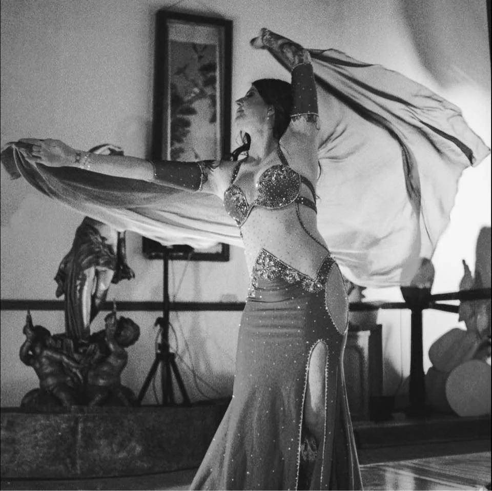 A woman in a belly dancing costume performing with a veil in a room decorated with artwork and sculptures.