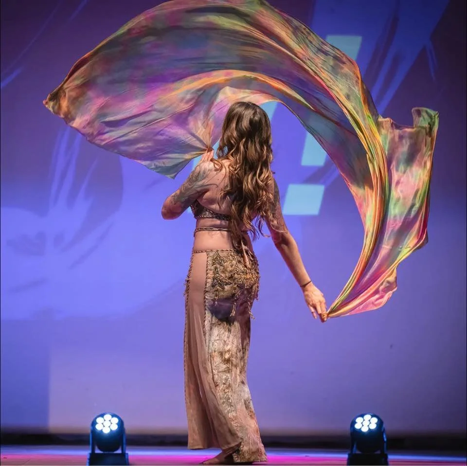 A woman with long wavy hair is dancing on stage, holding a colorful, flowing fabric in the air. She is wearing a patterned outfit and appears to be performing in front of a blue backdrop with stage lights.