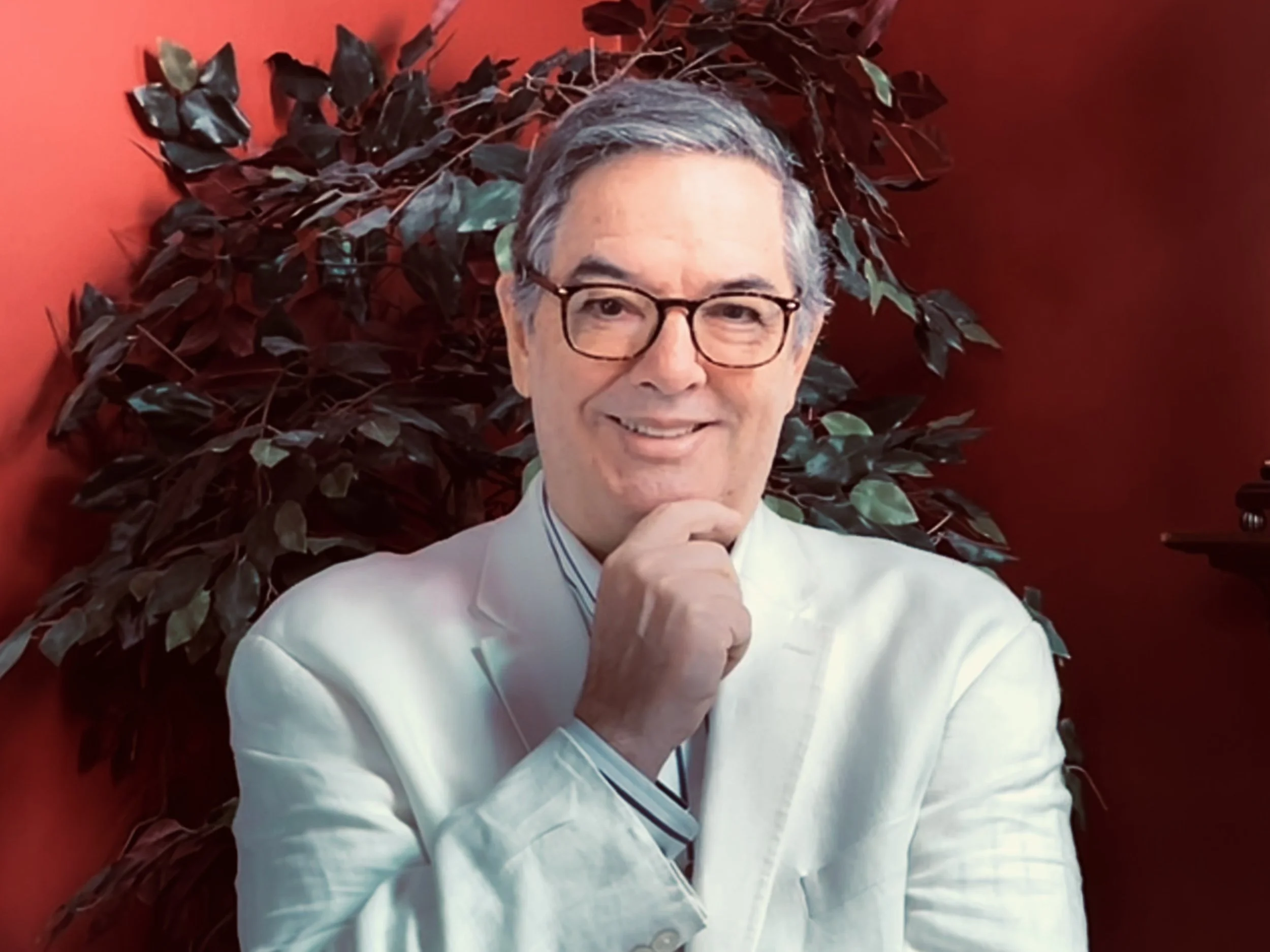 Author Thomas Maier posed for a picture wearing a white blazer with a striped button down and glasses. His right hand is holding his chin and he is in front of a tall green artificial plant