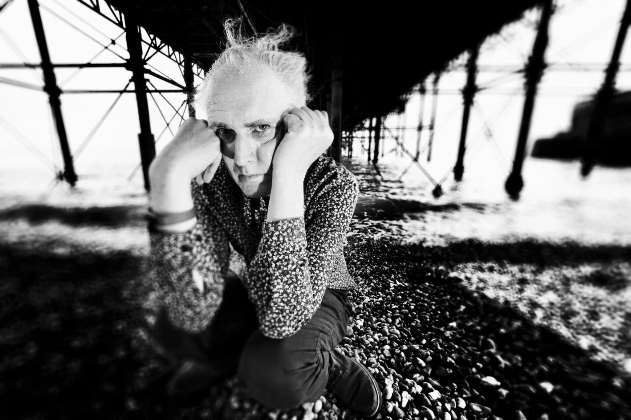 A woman with light-colored hair and a patterned shirt rests her head and hands on her knees on a pebble beach under a pier, with water and structural supports in the background. The image is in black and white.
