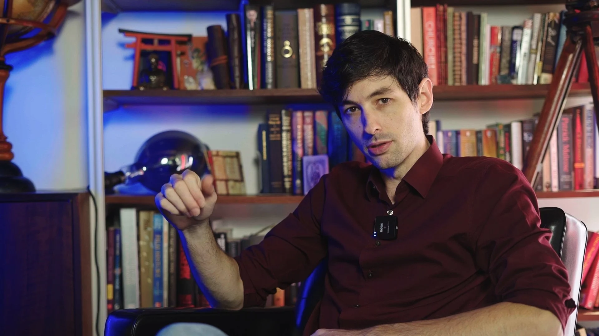 A young man with dark hair wearing a maroon shirt is seated in front of a bookshelf filled with books. He is holding his fist up, and a small black microphone is clipped to his shirt. The background includes a globe, a small red torii gate statue, and various decorative items, with a mix of natural and blue lighting.