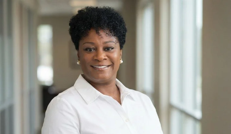 A smiling African American woman with short curly hair, wearing a white blouse, standing in a well-lit indoor space with large windows.