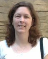Photo of a woman with wavy brown hair, smiling, wearing a white top, standing outdoors in front of a brick wall.
