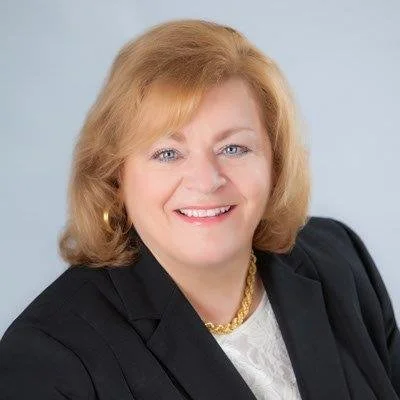 A professional woman with blonde hair wearing a black blazer, white blouse, and gold jewelry, smiling in a studio portrait.