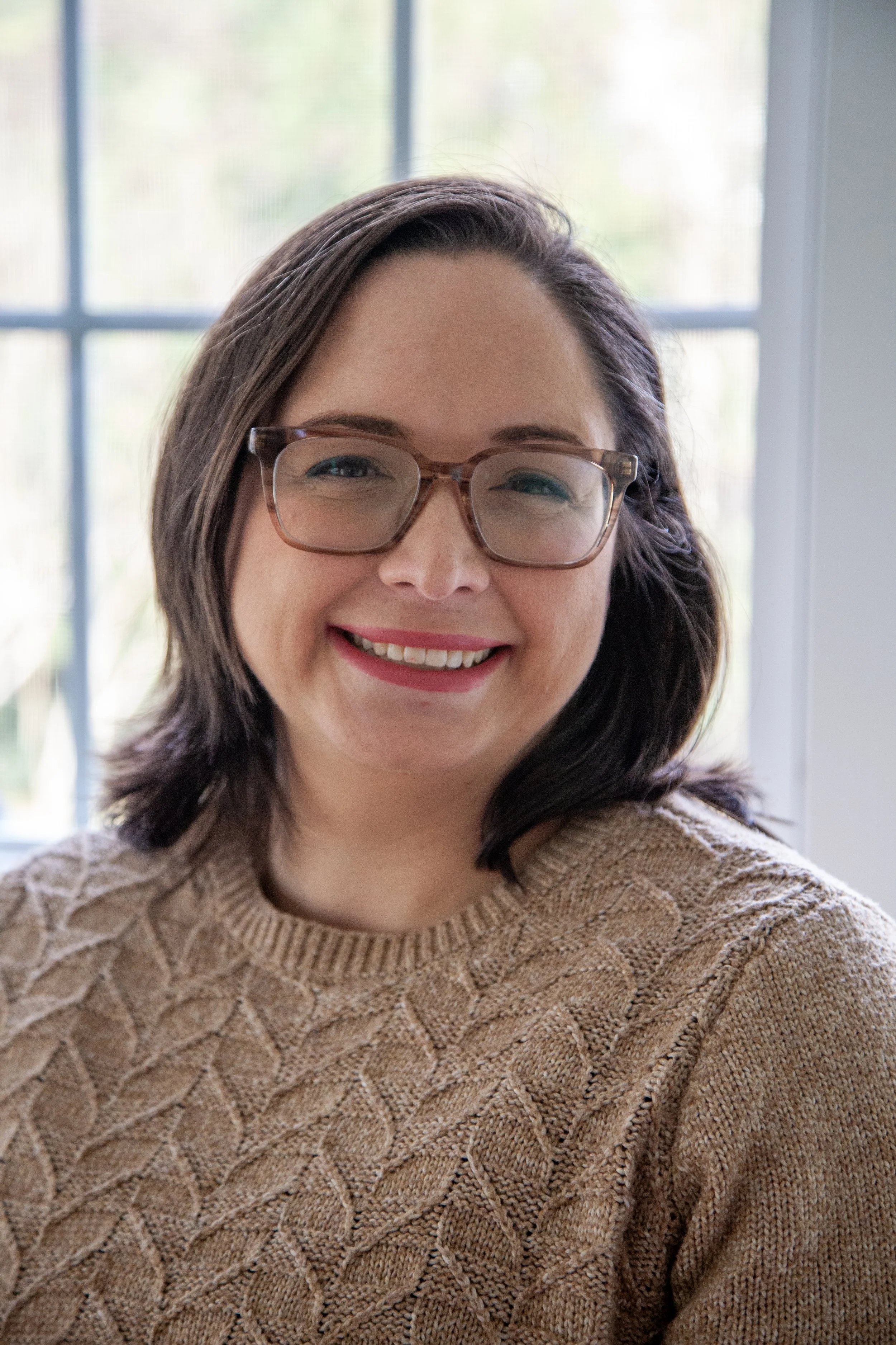 A woman with shoulder-length dark hair, glasses, and red lipstick smiling, wearing a tan knitted sweater, sitting by a window with a blurred background of trees and sky.