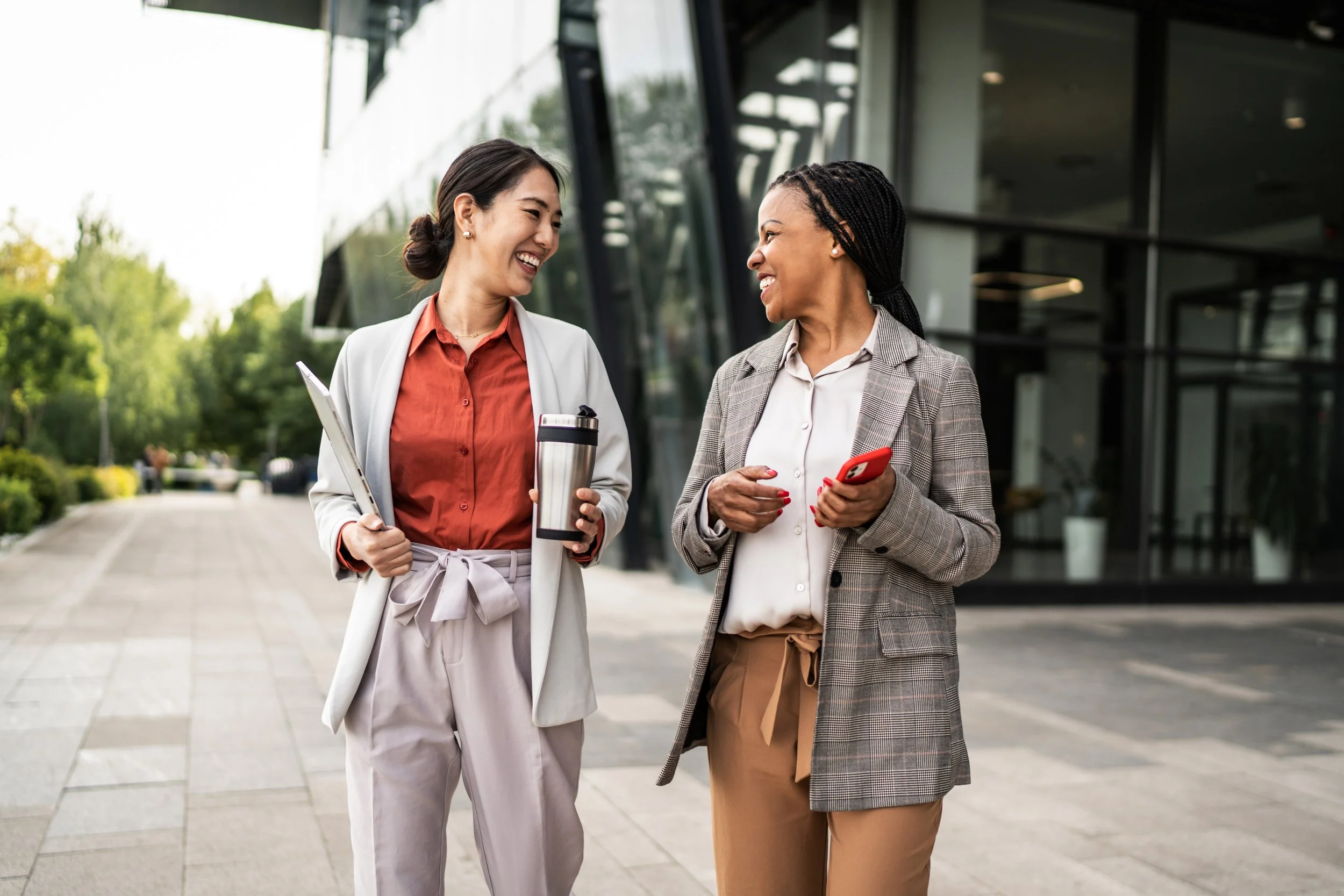 Two women are smiling and talking to each other outside a modern building with glass windows. One woman has dark braided hair and is holding a red phone, while the other has dark hair tied back and is holding a laptop and a travel mug.