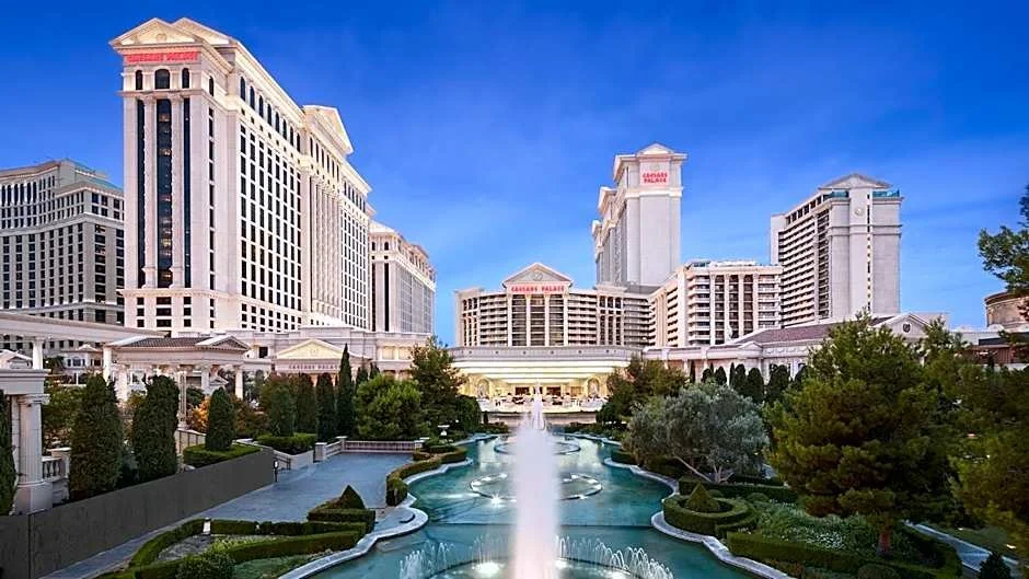 View of Caesars Palace hotel and casino in Las Vegas, with a fountain and landscaped gardens in the foreground under a clear blue sky.