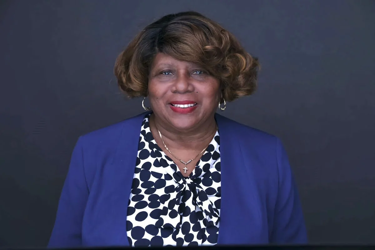 Portrait of an African American woman with short brown hair, smiling, wearing a blue blazer, polka dot blouse, and jewelry against a dark background.