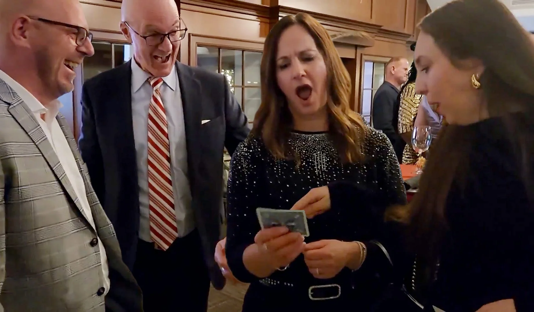 Group of four people at a social gathering, looking at a photo on a phone, with other guests in the background in a warmly lit room.