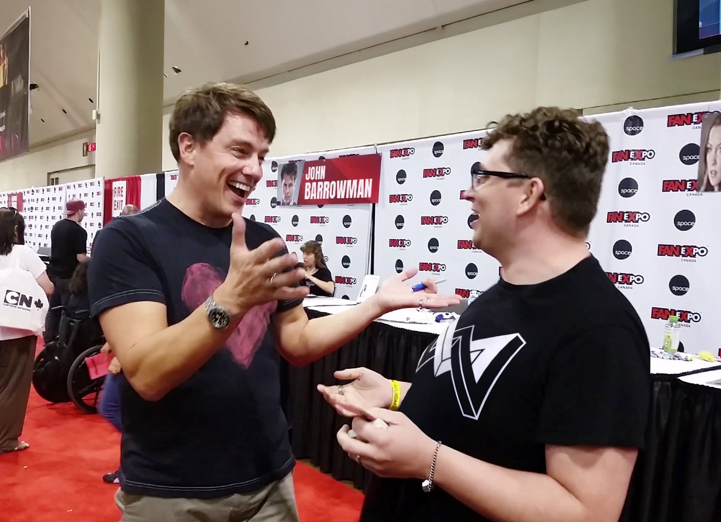 Two men engaging in conversation at Fan Expo event, with a backdrop displaying John Barrowman's name and photos, and other attendees in the background.