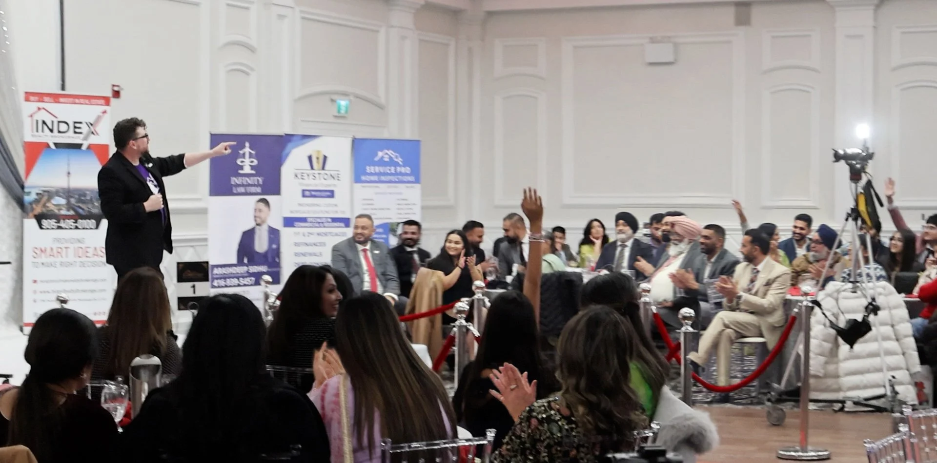 A speaker is giving a presentation at a conference or seminar with a diverse audience of men and women seated at tables. Some audience members are raising their hands, and others are clapping. The room has large banners with logos and promotional information along the back wall, and a camera is positioned on a tripod capturing the event.