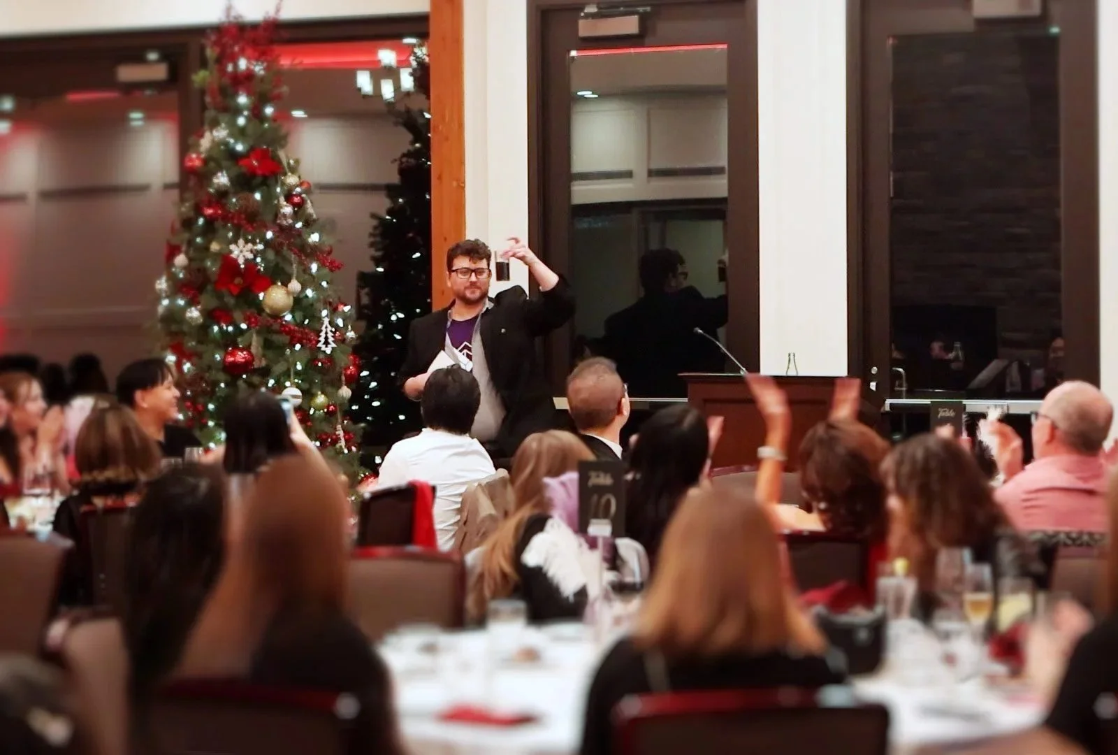 A man standing in front of a Christmas tree, holding a paper and speaking to an audience in a banquet hall.