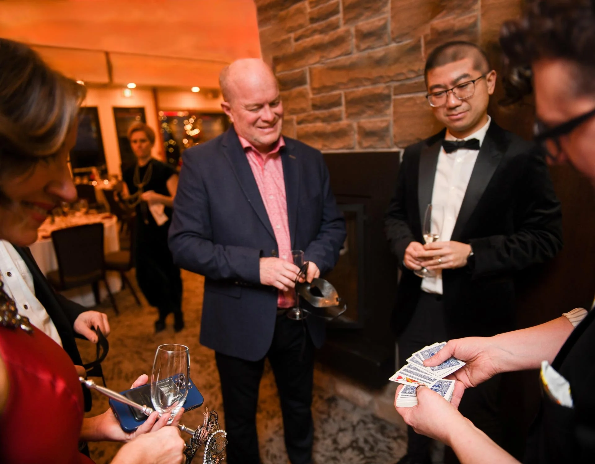 People at a social gathering playing cards and holding glasses of wine in a warmly lit room with brick walls.