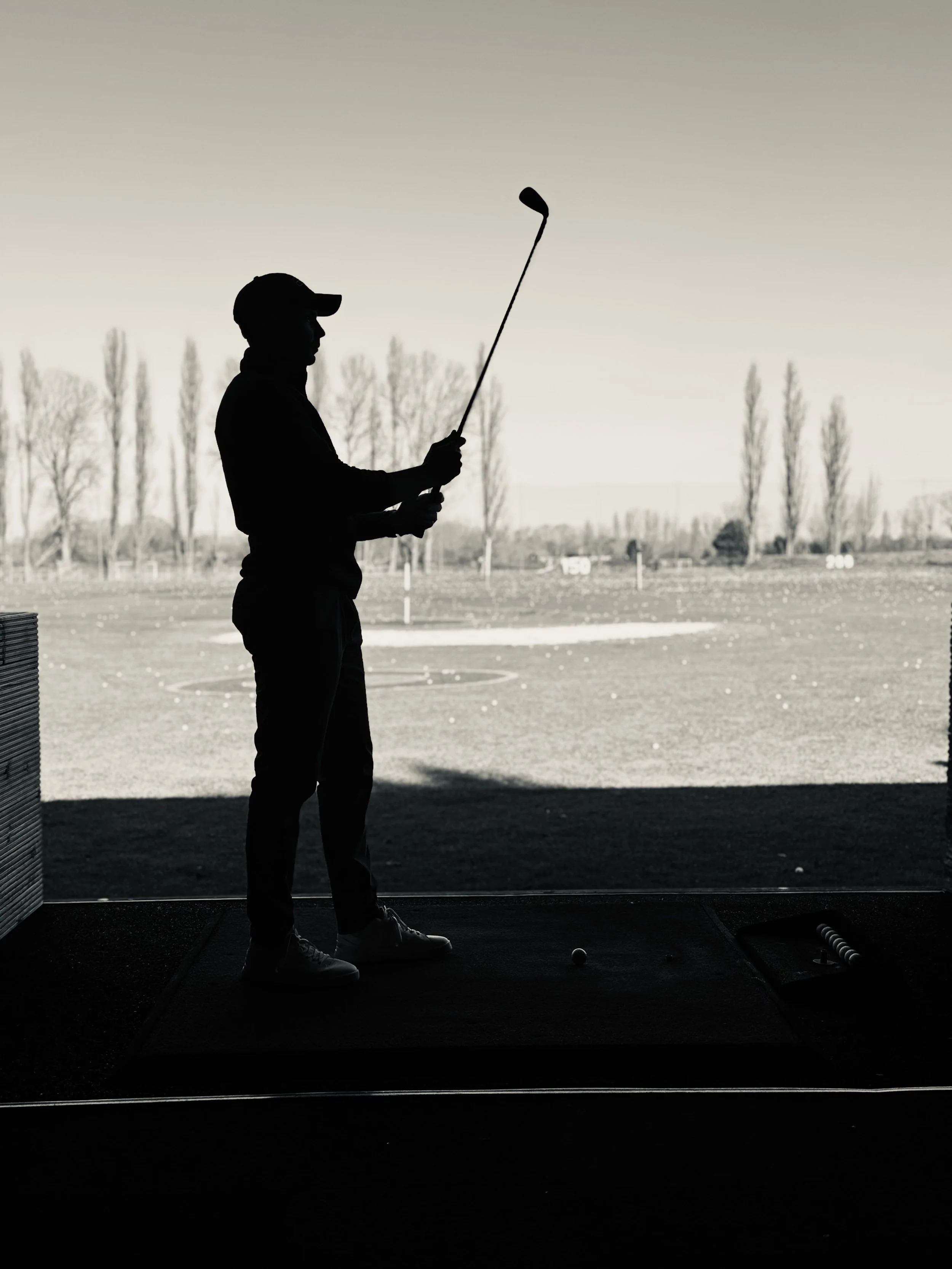Silhouette of a man in a cap holding a golf club, standing on a golf tee at a golf driving range with trees and open field in the background.