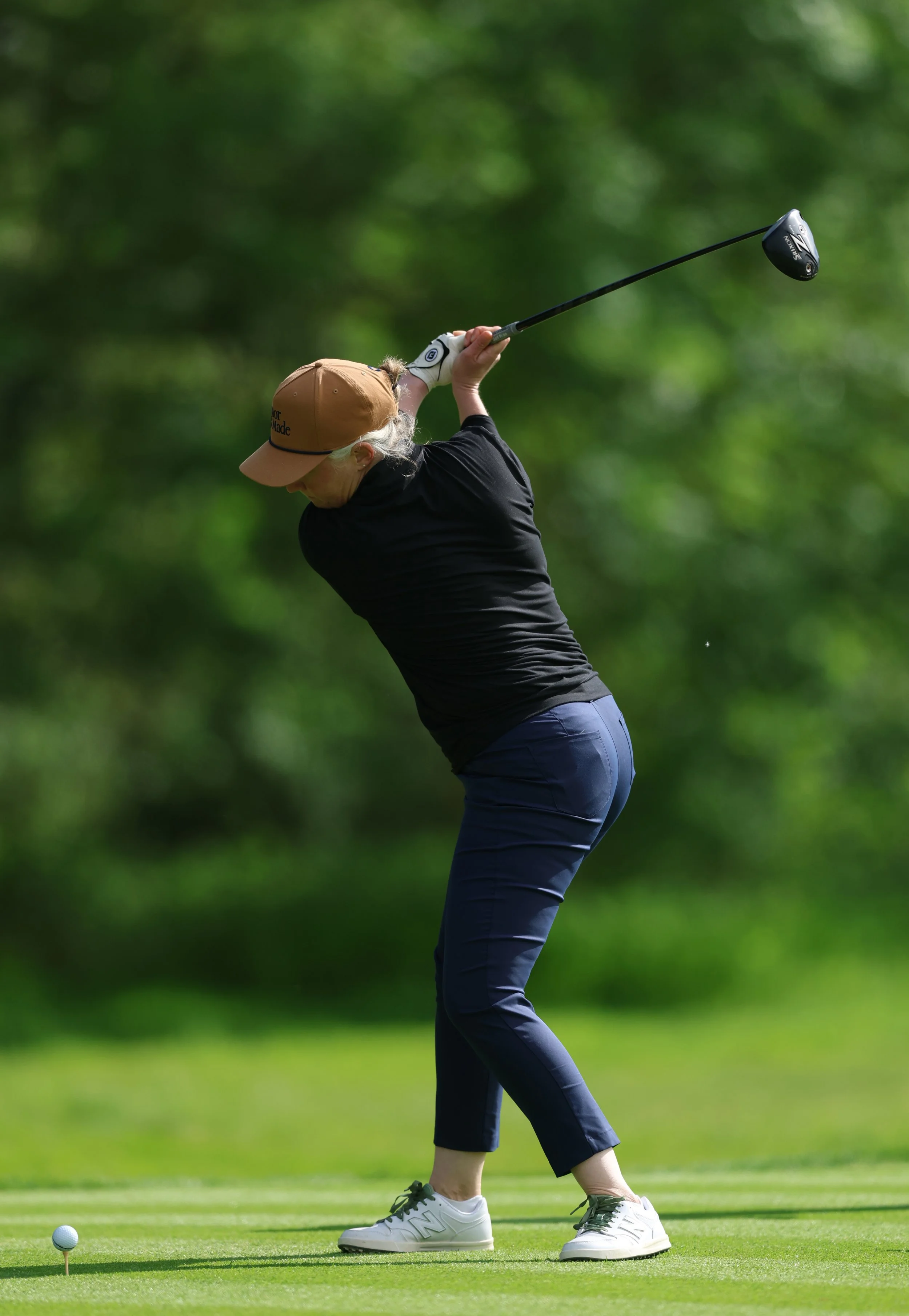 A woman in a black shirt, blue pants, and a tan cap is taking a golf shot on a green golf course. She is holding a golf club and focusing on a golf ball on a tee.