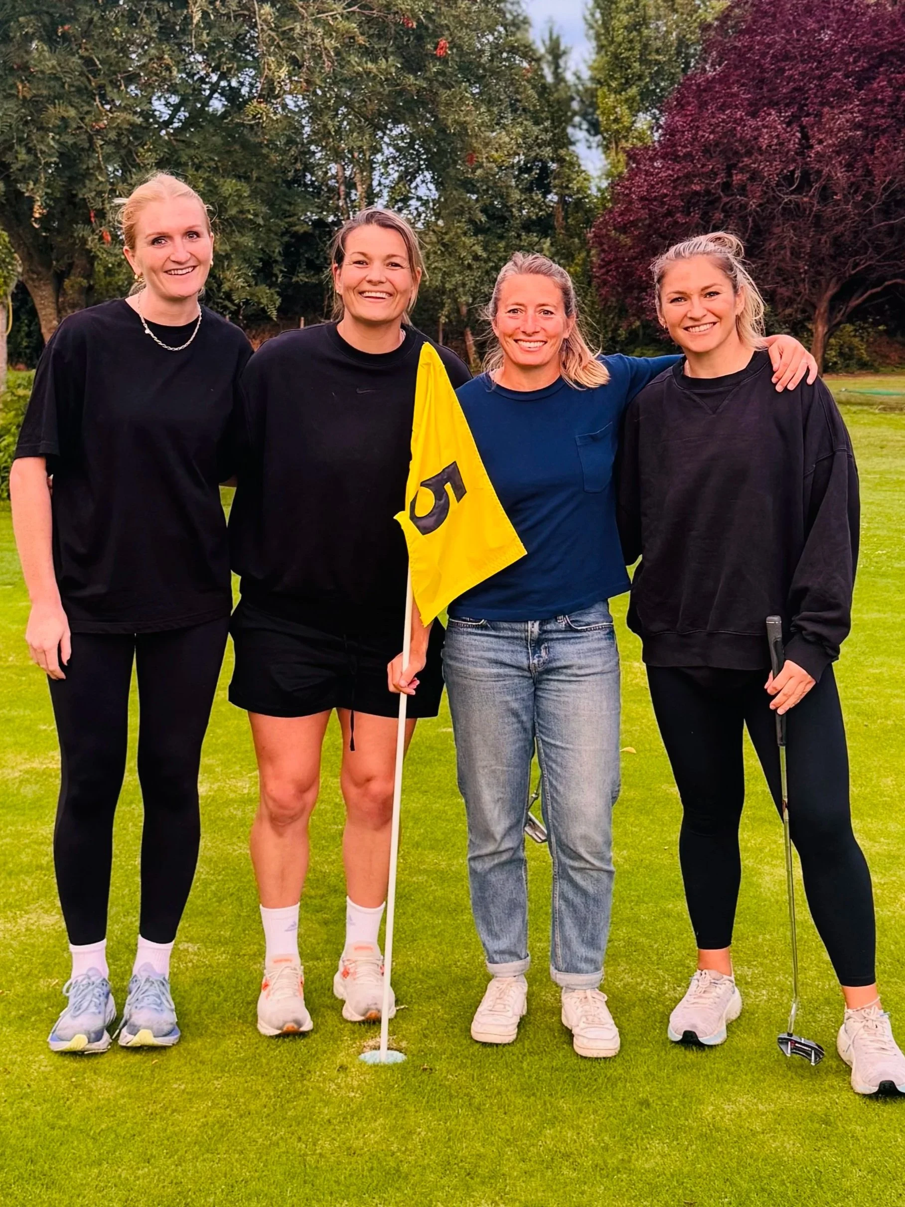 Four women standing on a golf course, smiling at the camera. The woman second from the left is holding a yellow flag with the number 5. All are dressed in casual sportswear, and the background features trees and grass.