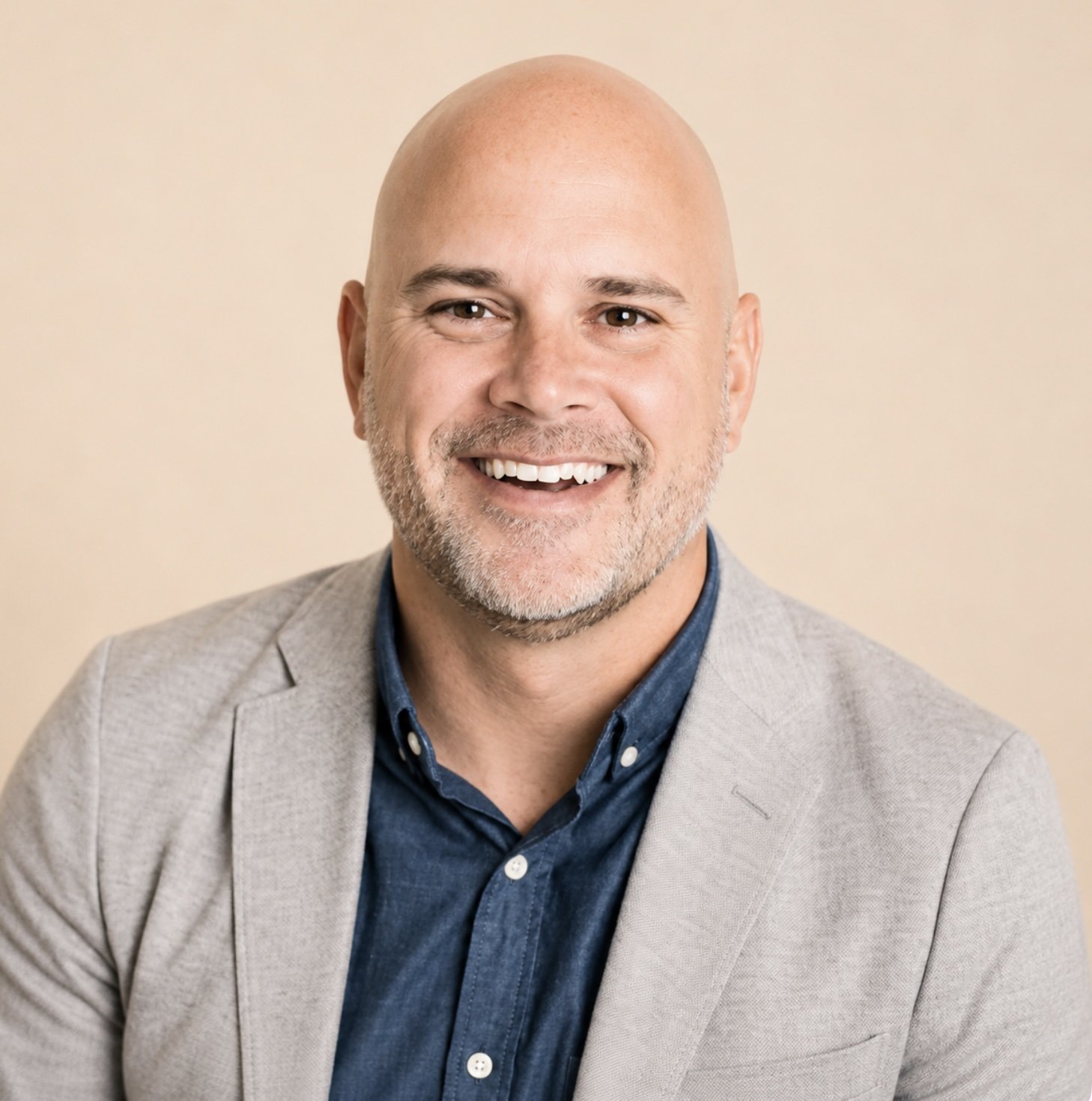 A smiling bald man with a beard, wearing a light gray blazer over a dark blue shirt, shot in front of a beige background.