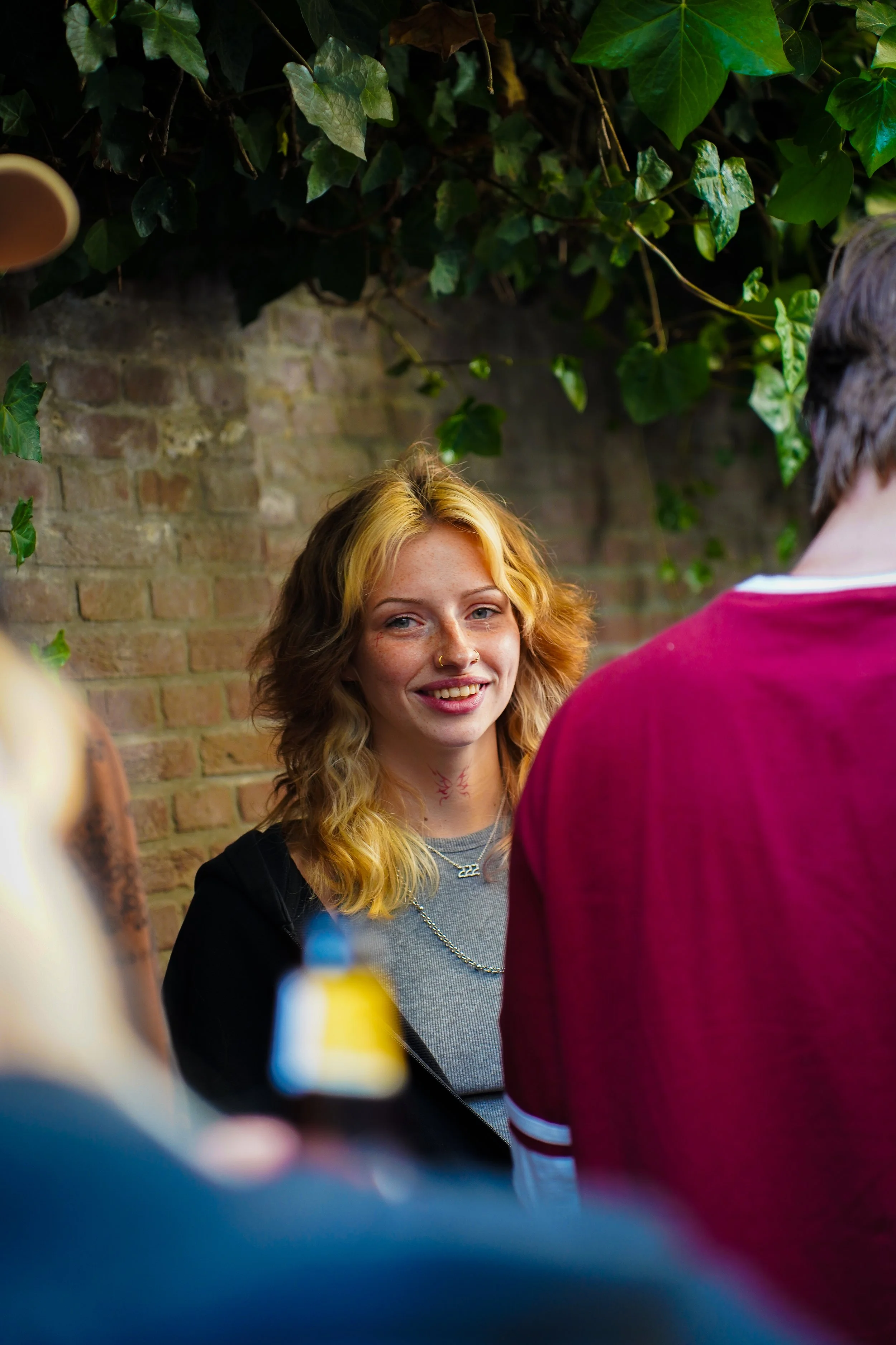A young woman with wavy blonde hair, wearing a grey shirt and black hoodie, smiling and talking to a group in an outdoor setting with a brick wall and greenery in the background.