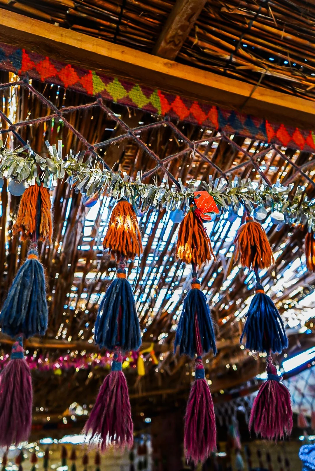 Colorful tassels hanging from a string in a rustic, thatched-roof setting with colorful decorations.