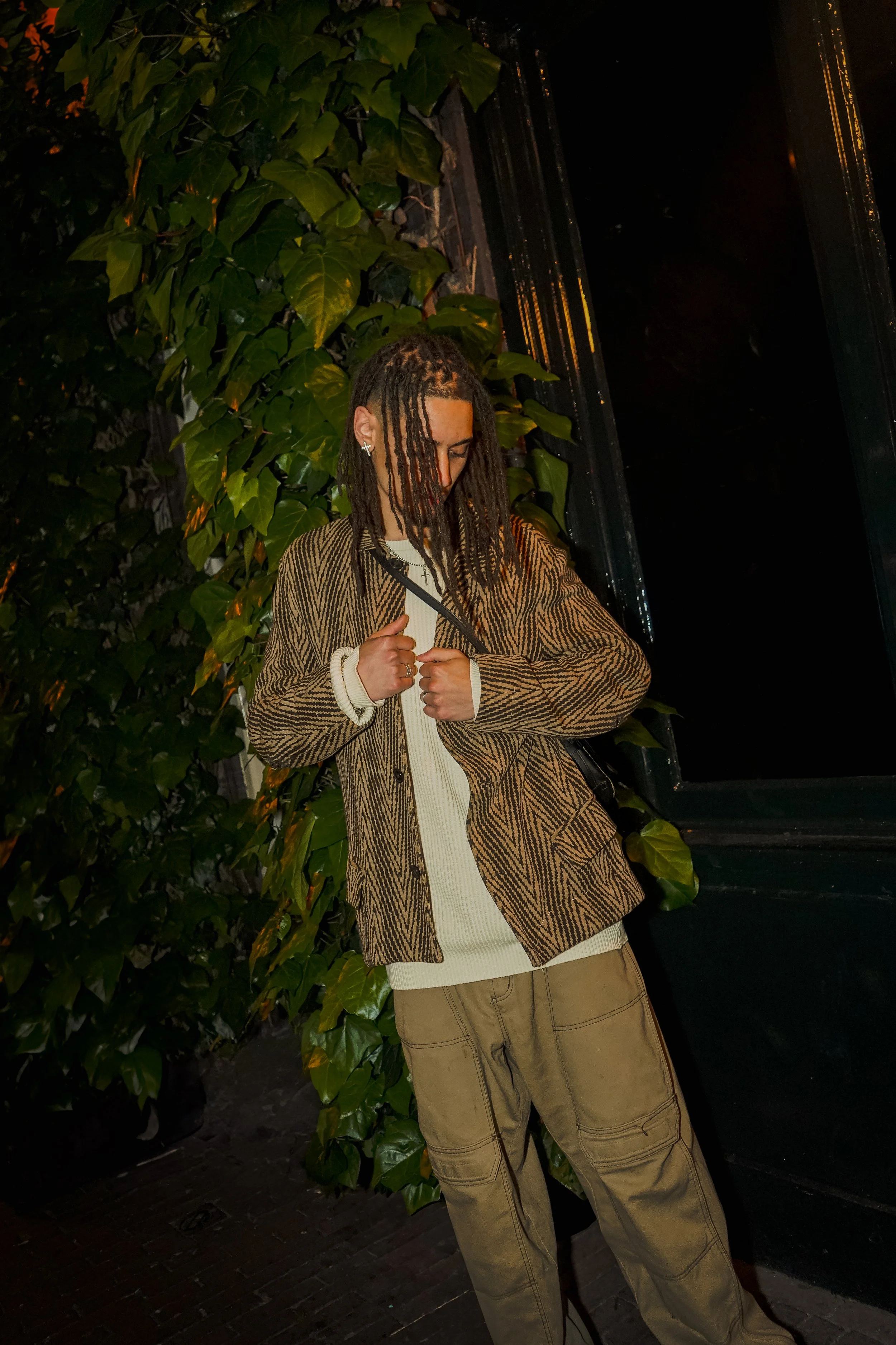 A person with long dreadlocks wearing a patterned jacket, cream sweater, and cargo pants standing outdoors at night near green leafy plants and a black structure.