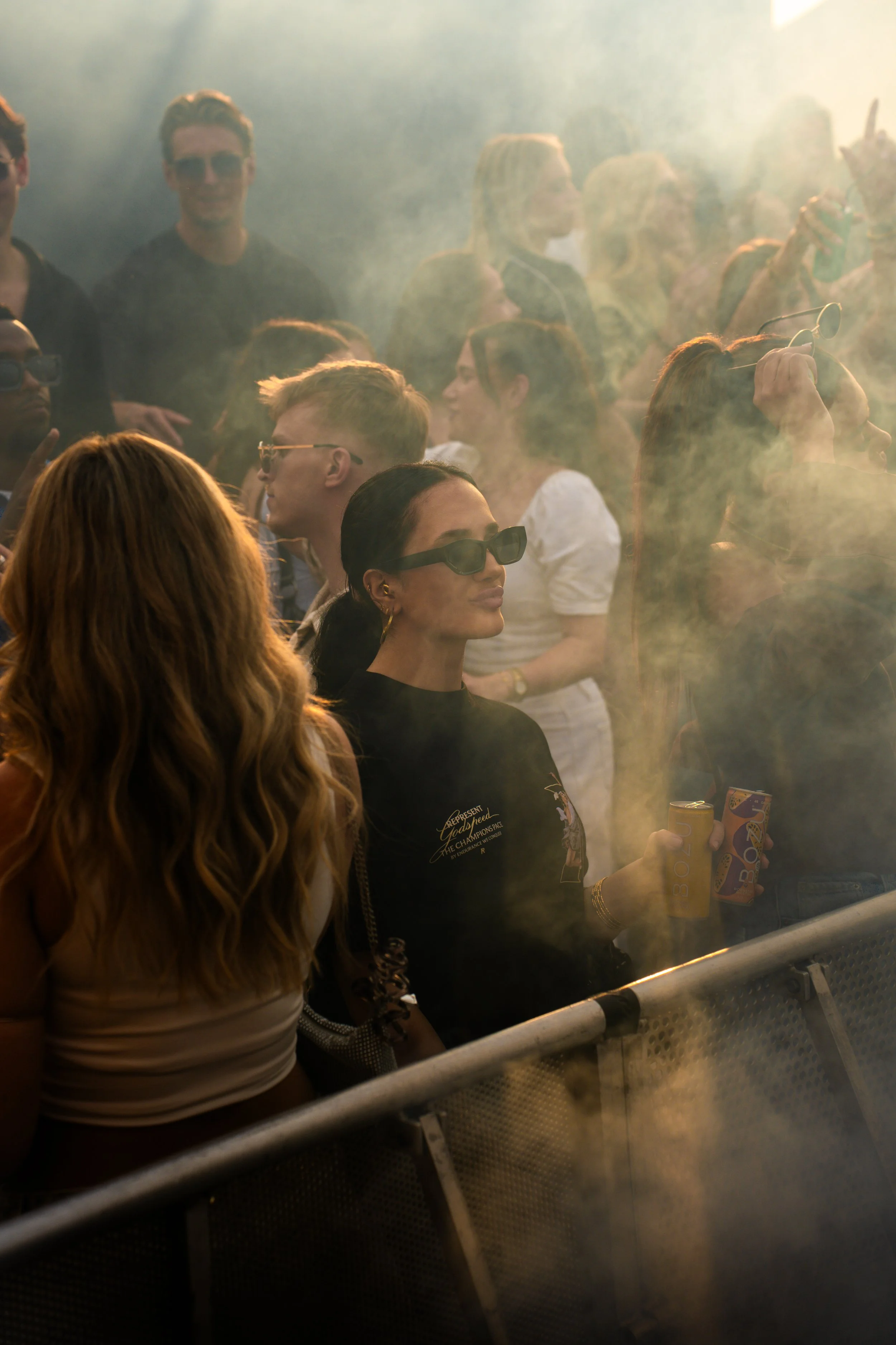 Crowd of people at an outdoor event, some wearing sunglasses, standing behind a metal barricade, with smoke or fog in the air.